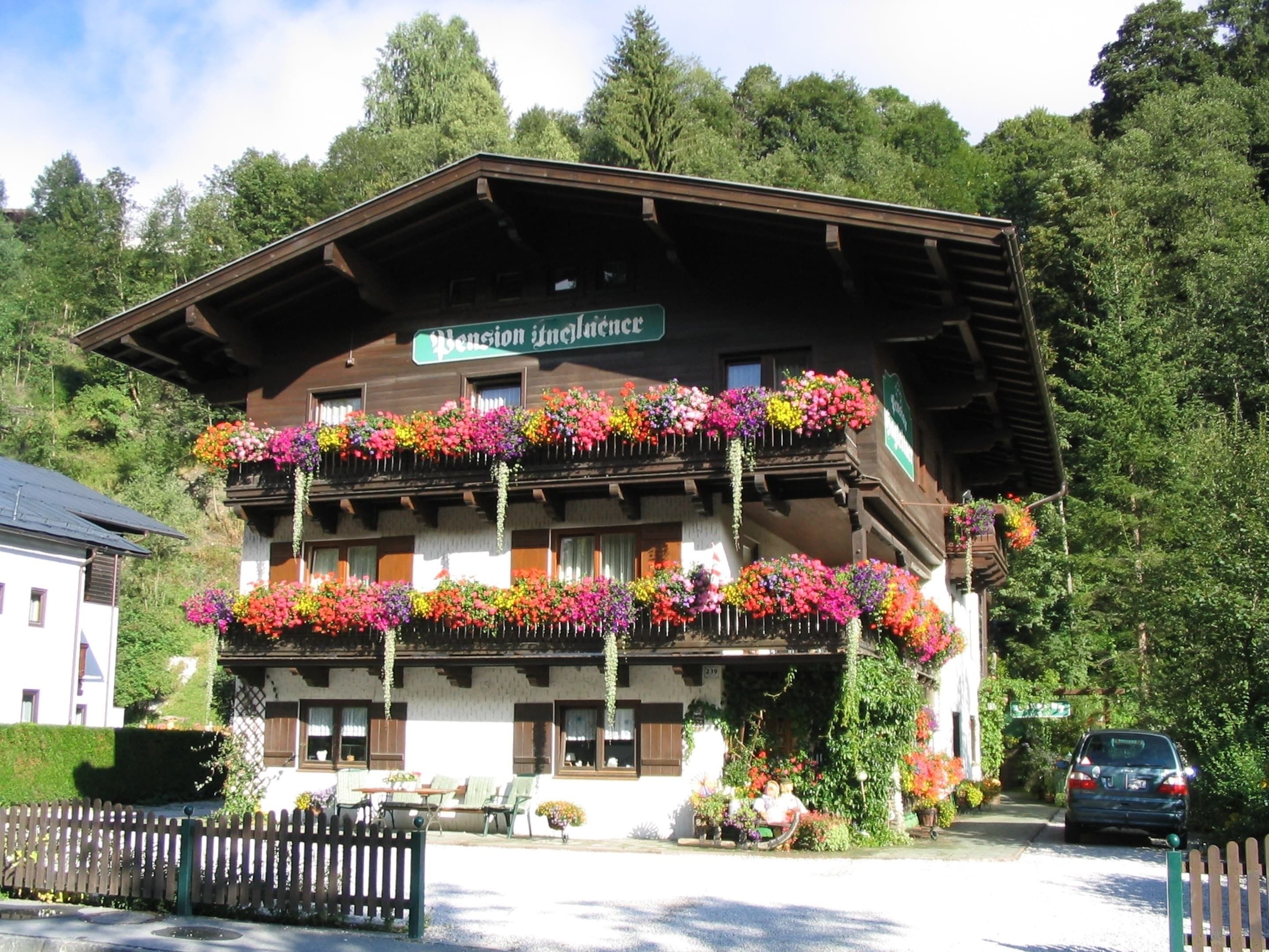 A traditional chalet with multiple balconies adorned with colorful flowers, a sign indicating it is a pension, and a car parked in front.