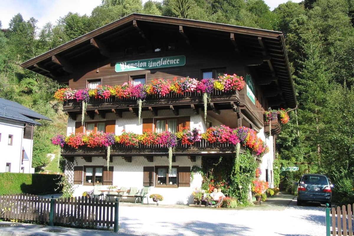 A traditional chalet with multiple balconies adorned with colorful flowers, a sign indicating it is a pension, and a car parked in front.