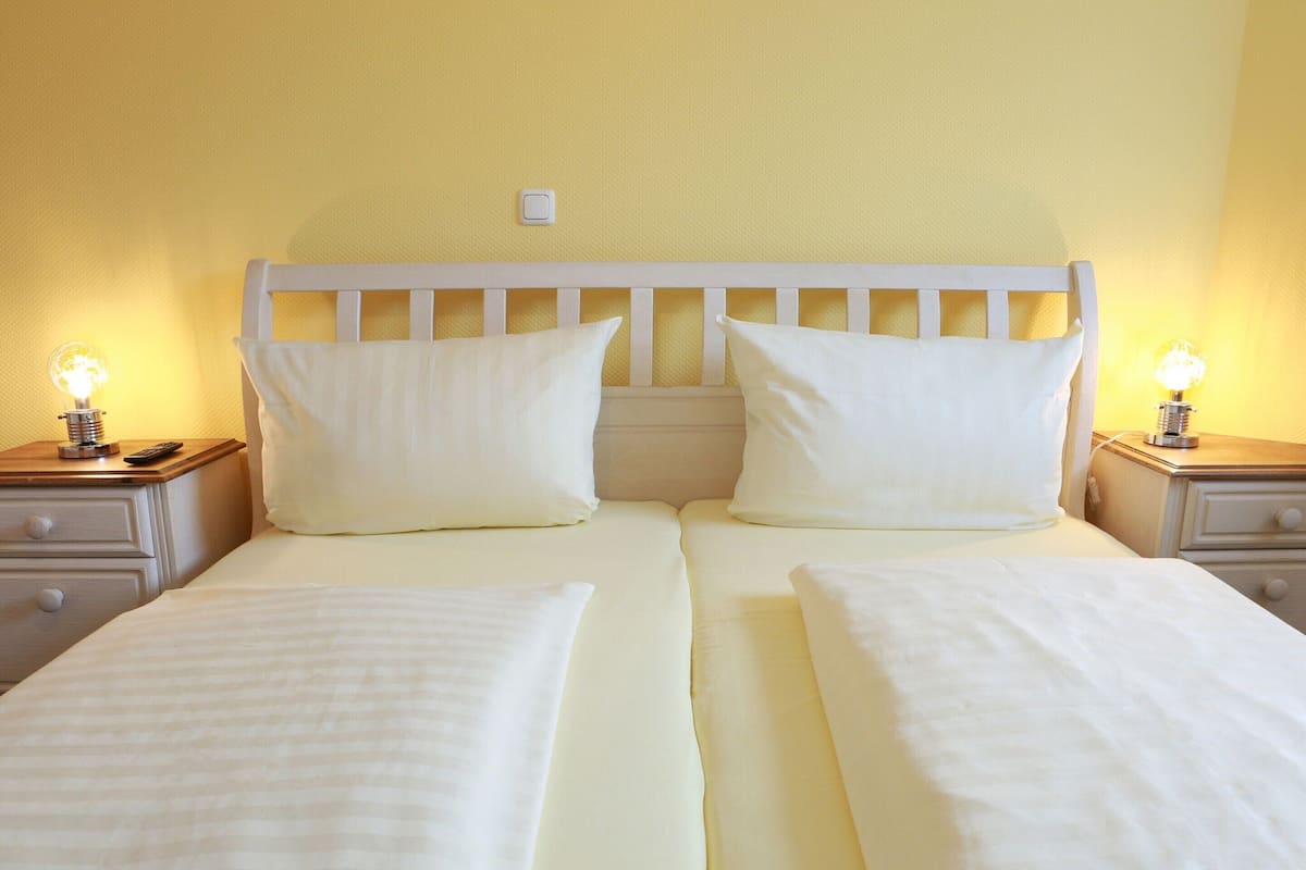 Un letto matrimoniale con biancheria da letto bianca, due comodini con lampade e un interruttore a muro sopra il letto.