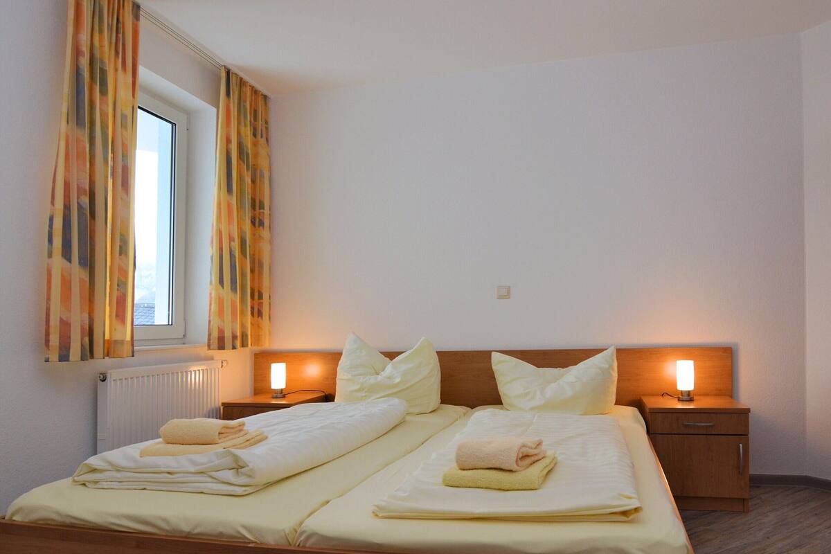 Un letto matrimoniale con due cuscini, un comodino con una lampada e una finestra con tende.