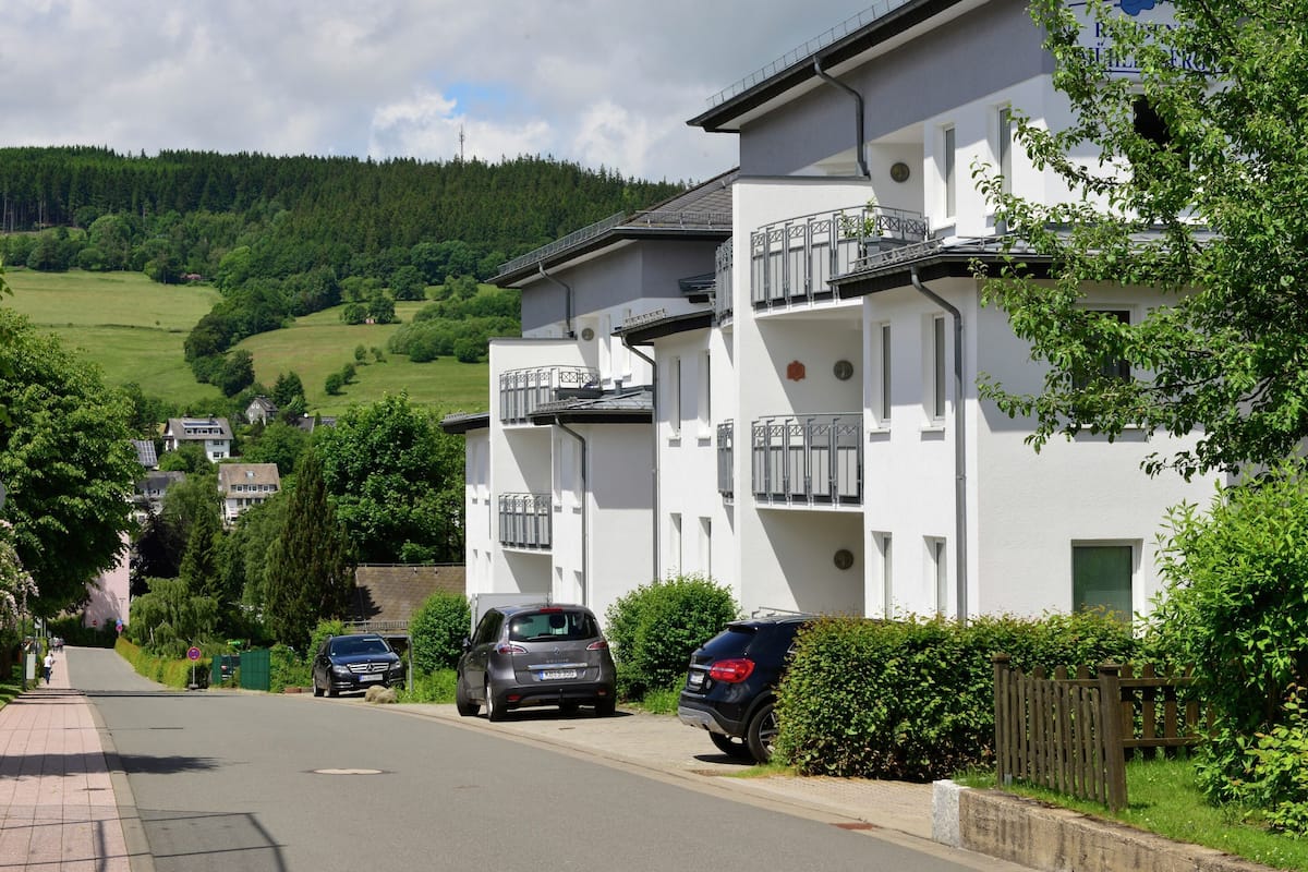 A residential building with balconies, a paved street, and a hilly landscape in the background.