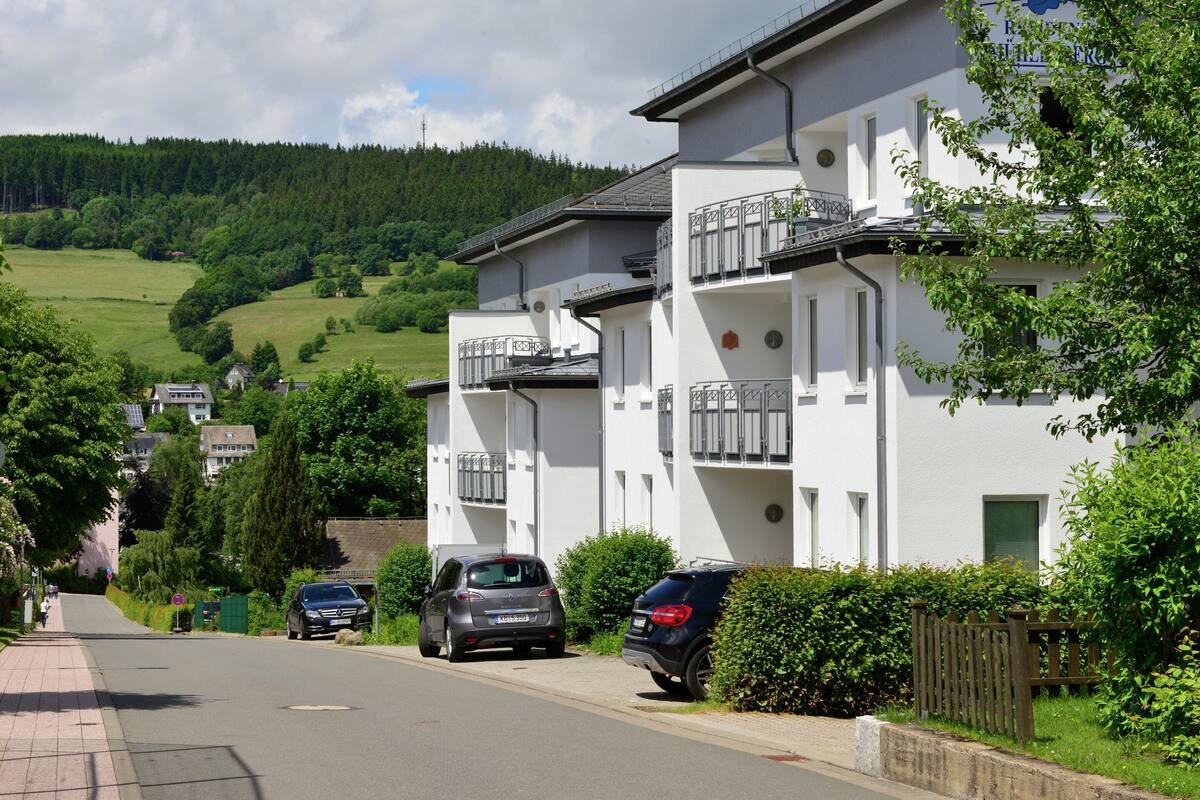 Ein Wohngebäude mit Balkonen, eine gepflasterte Straße und eine hügelige Landschaft im Hintergrund.