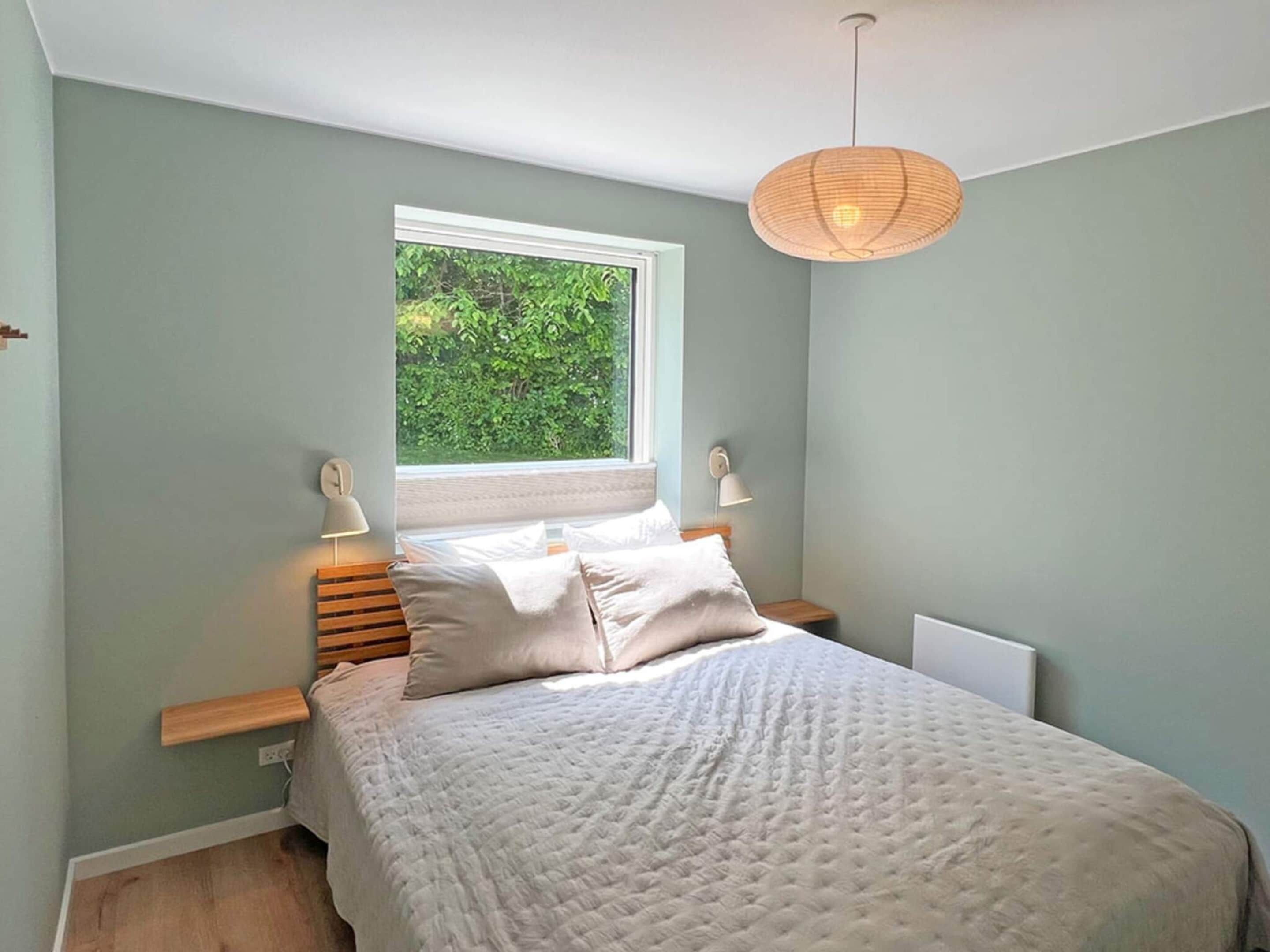 A bedroom with a bed, bedside lamps, a window with a view of greenery, and a hanging light fixture.