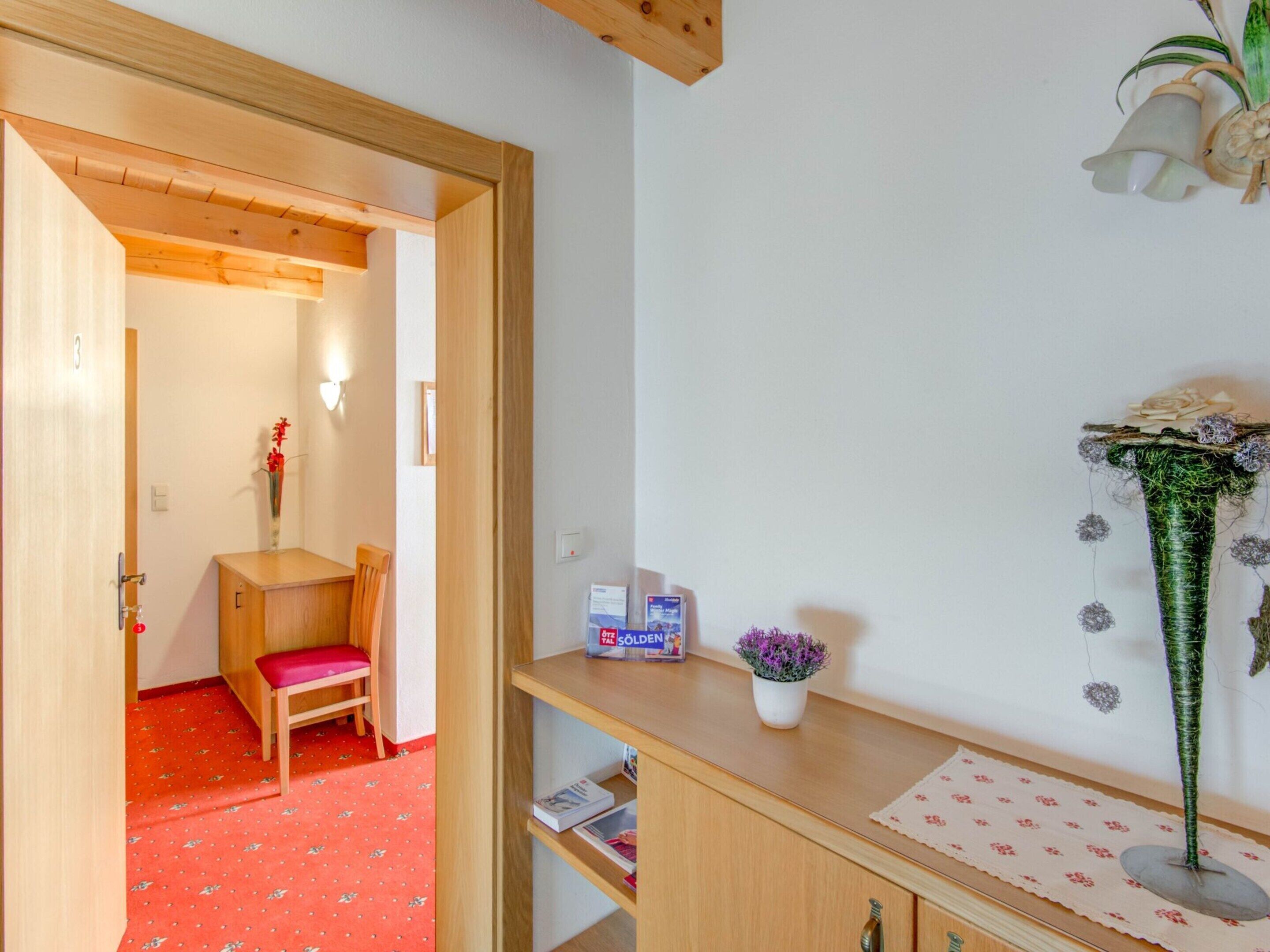 Una habitación con un banco de madera, una mesita con un reloj y un florero, y una planta decorativa en la pared.