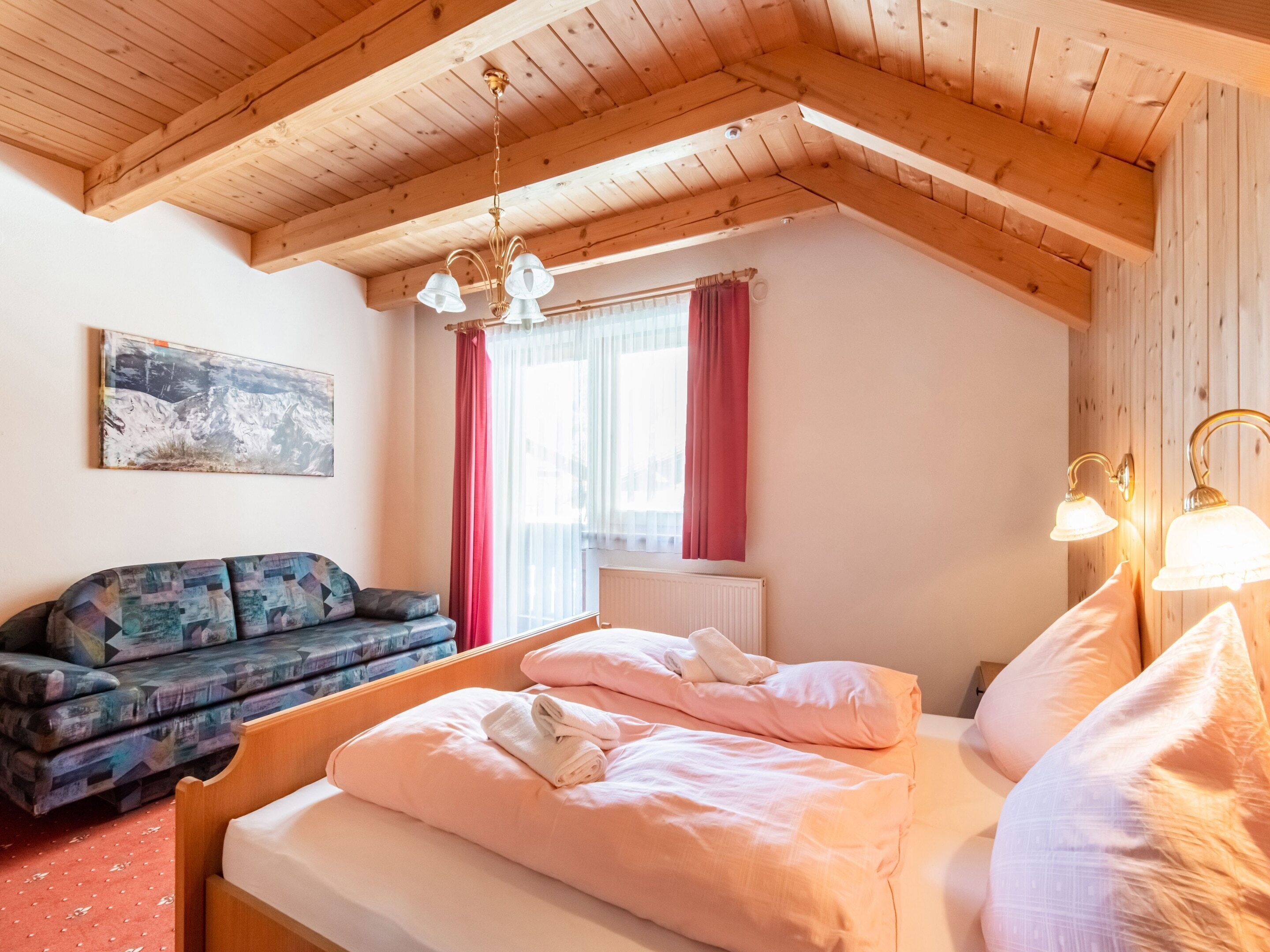 A cozy room with a wooden ceiling, a bed with pink bedding, a sofa with patterned cushions, and a window with red curtains.