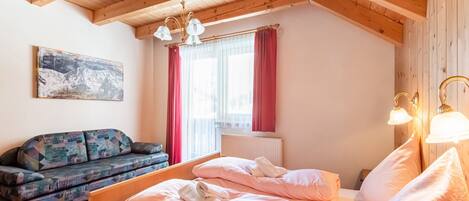 A cozy room with a wooden ceiling, a bed with pink bedding, a sofa with patterned cushions, and a window with red curtains.
