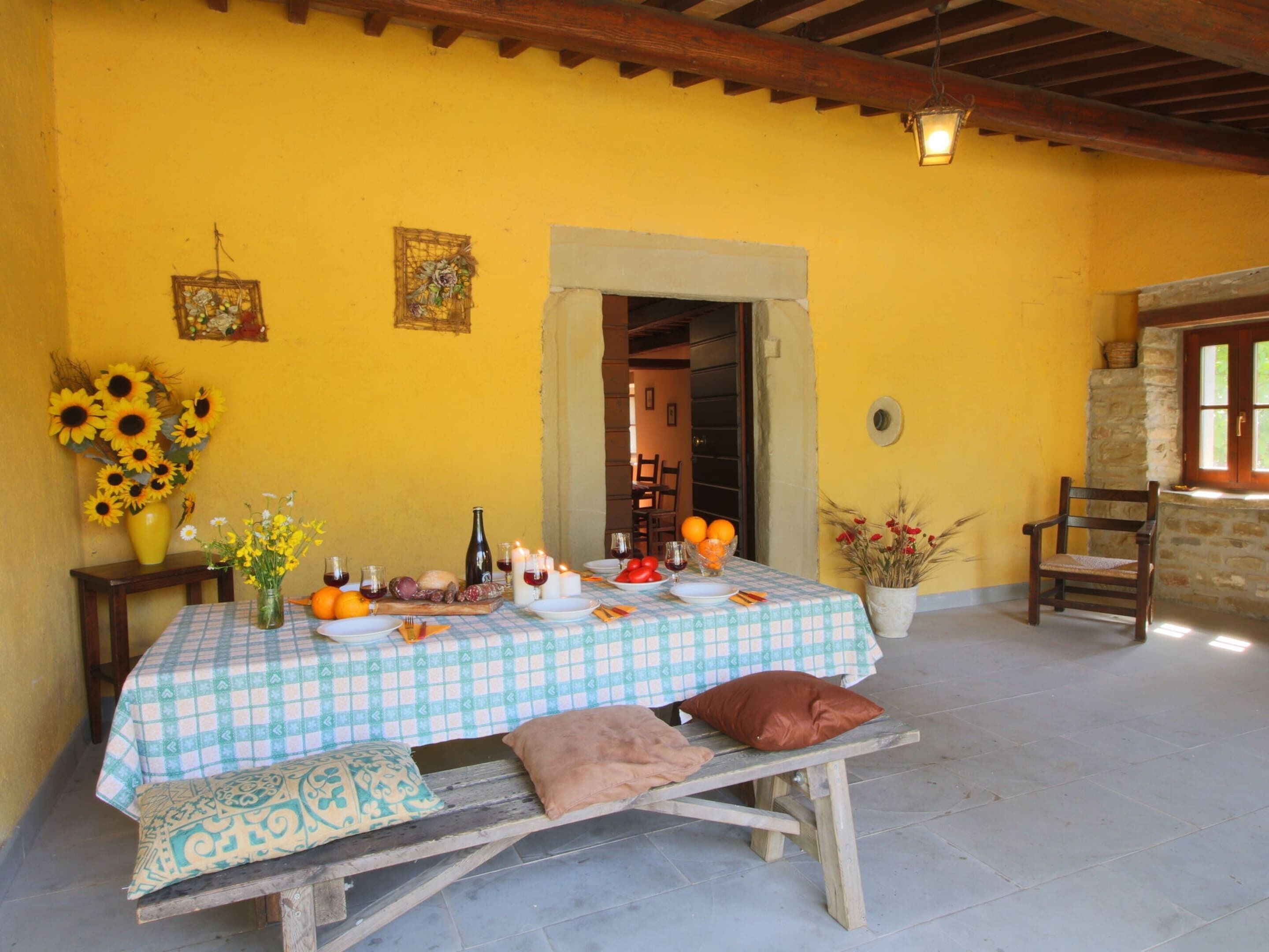 Un coin repas avec une nappe à carreaux, un banc et une chaise. On peut voir des fleurs, une bouteille et un panier sur la table.