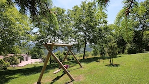 A wooden swing set in a grassy park area with trees and a building in the background. - Farmhouse in Apecchio With Swimming Pool, Terrace, Garden (Apecchio)