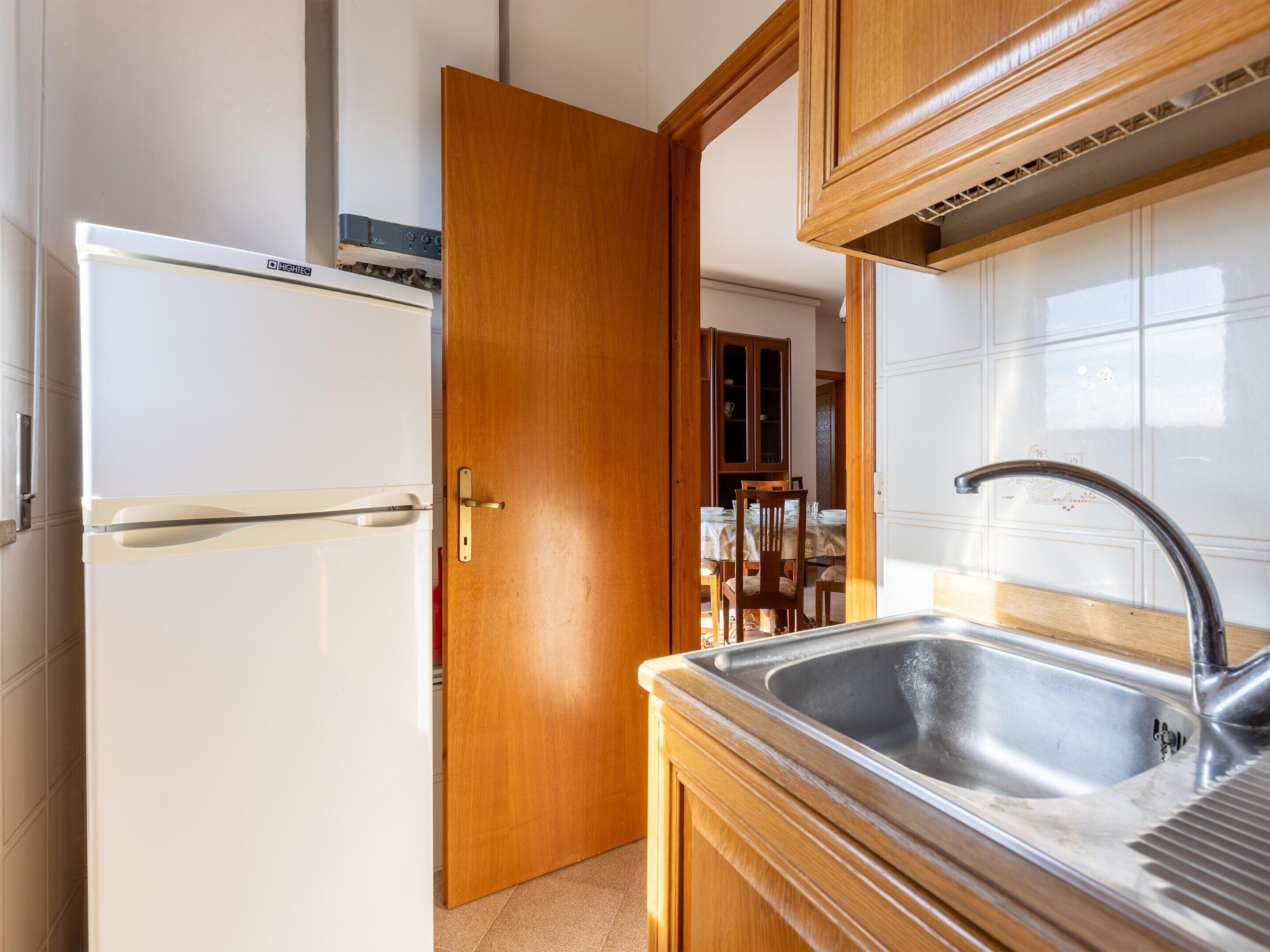 A kitchen with a white refrigerator, wooden cabinets, a stainless steel sink, and a dining area visible through an open door.