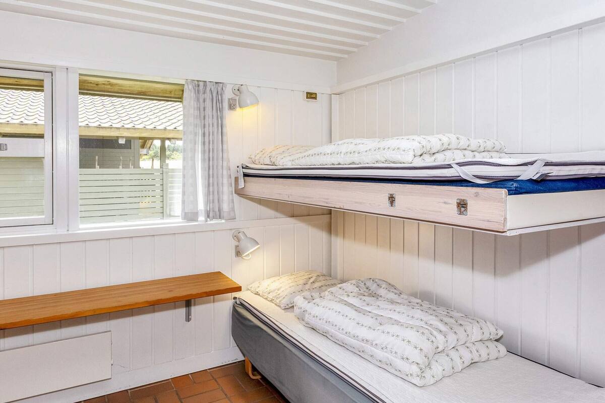 A dormitory room with bunk beds, a window with curtains, a bedside table, and a wall-mounted lamp.