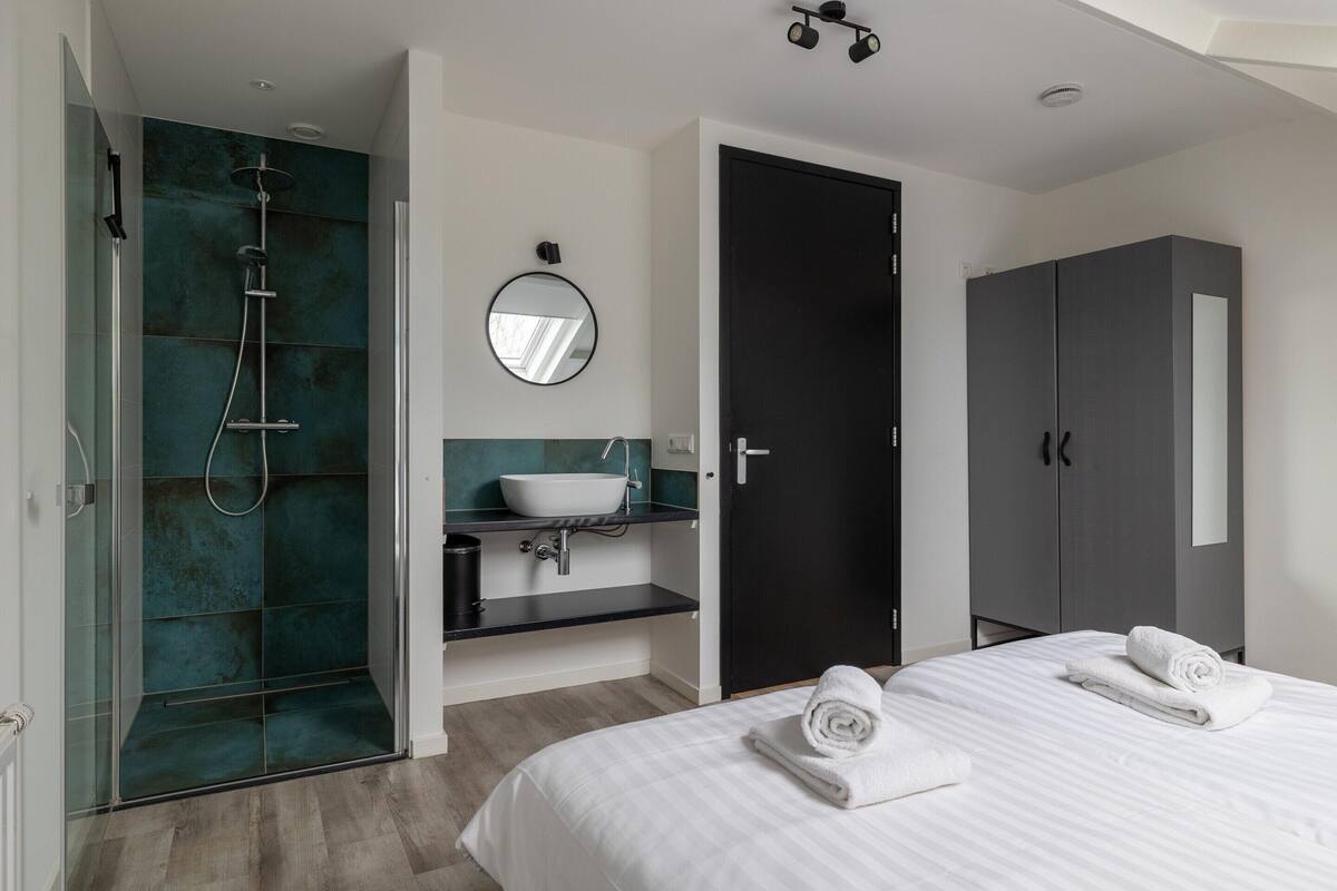Ein modernes Hotelzimmer mit einer gläsernen Dusche, einem weißen Bett mit ordentlich gefalteten Handtüchern, einem dunklen Kleiderschrank und einem runden Spiegel über dem Waschbecken.