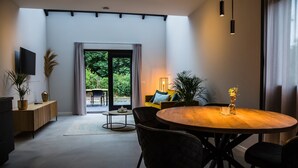 Ein modernes Wohnzimmer mit einem runden Holztisch, schwarzen Stühlen, einer Couch mit Kissen, einem an der Wand montierten Fernseher und einem großen Fenster mit Vorhängen.
