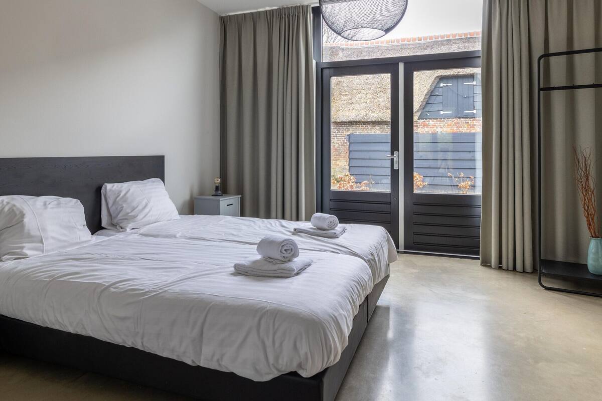 A modern bedroom with a large bed, two bedside tables, a nightstand, and a view through a sliding glass door to the outside.