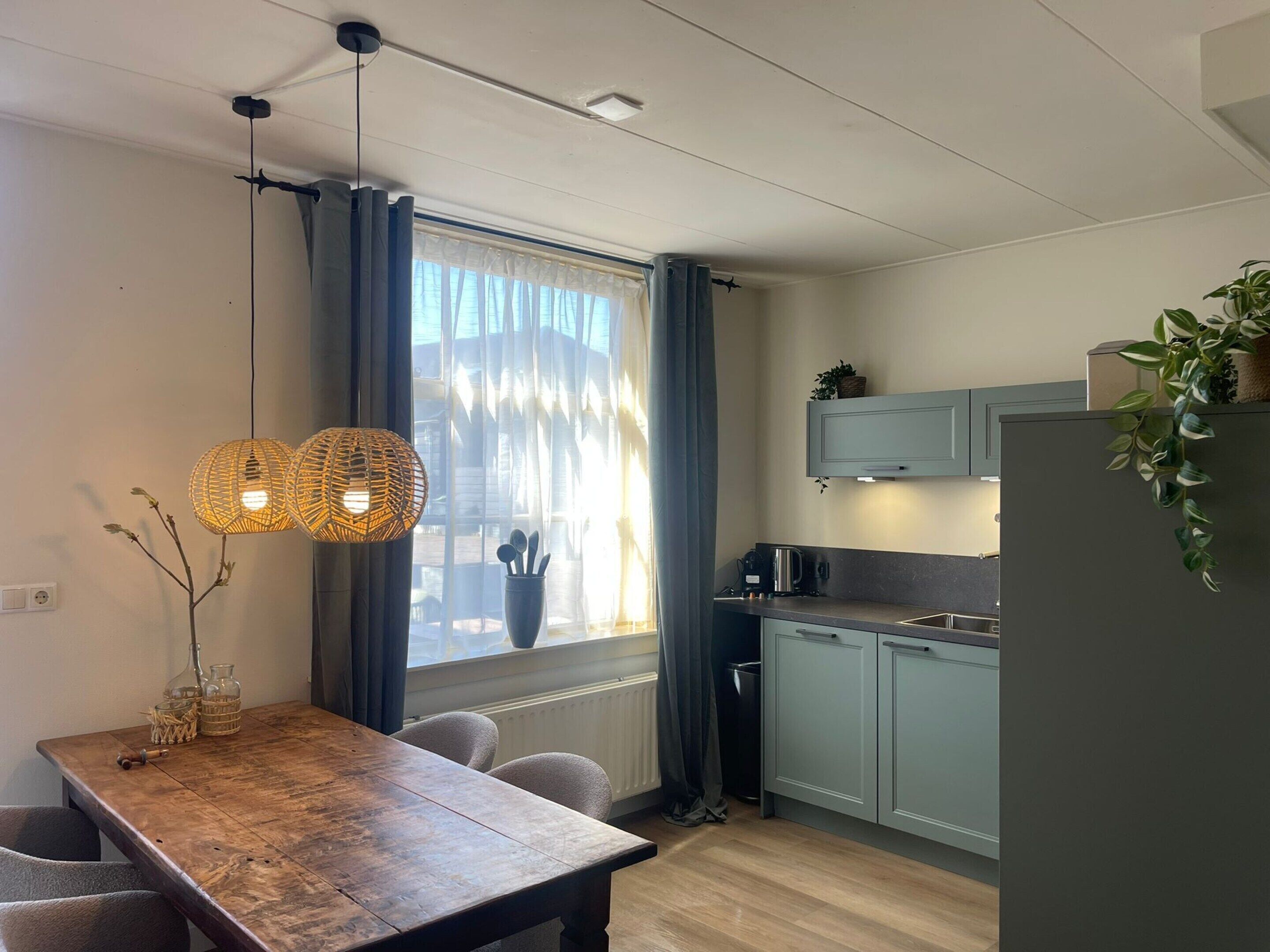 Et kompakt køkken med spiseplads, der indeholder et træbord, stole, en hængende lampe og et vindue med gardiner.