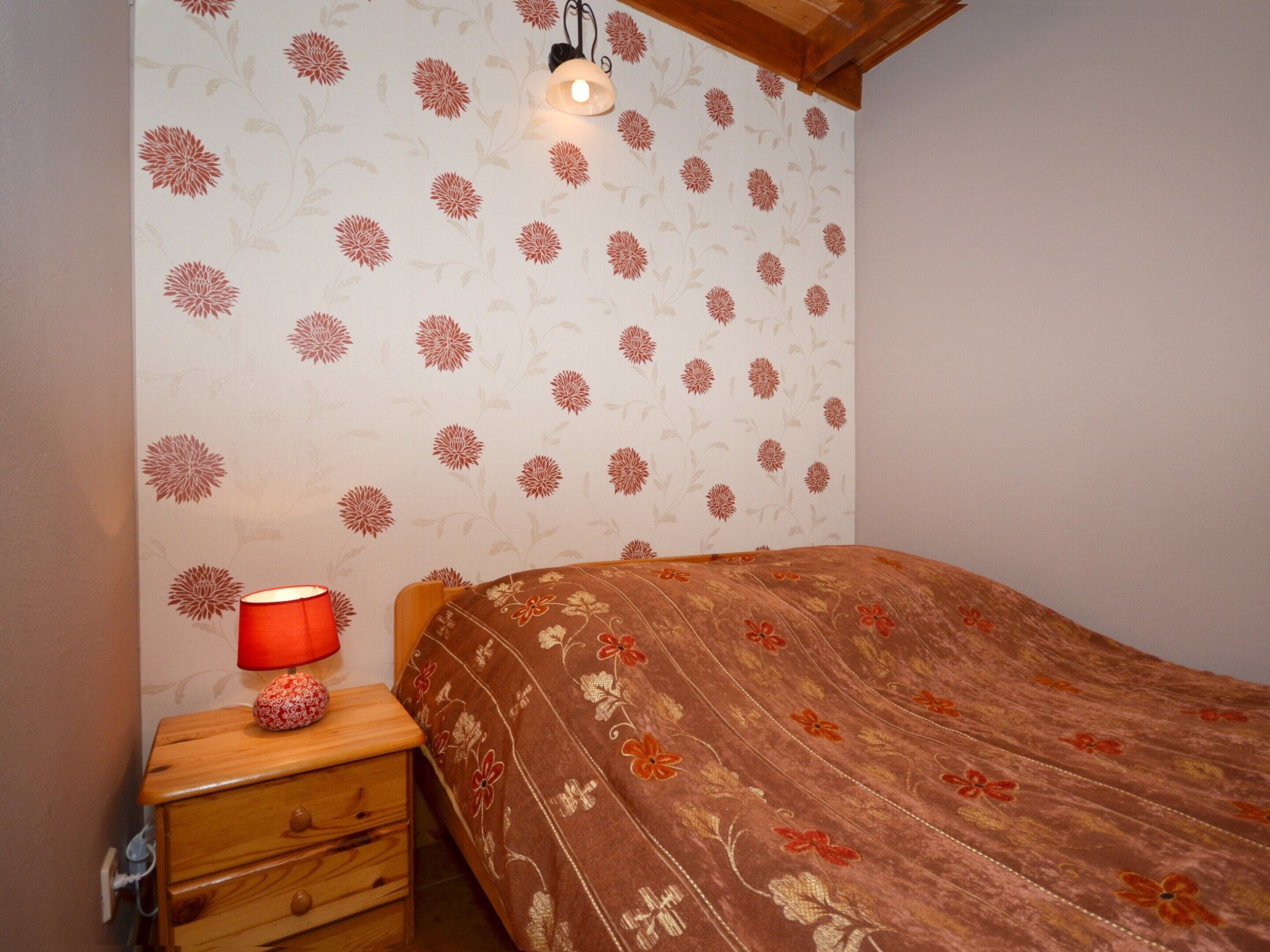 Une chambre à coucher avec un papier peint à motifs floraux, une table de chevet en bois avec une lampe et un lit recouvert d’une couvre-lit à motifs floraux.