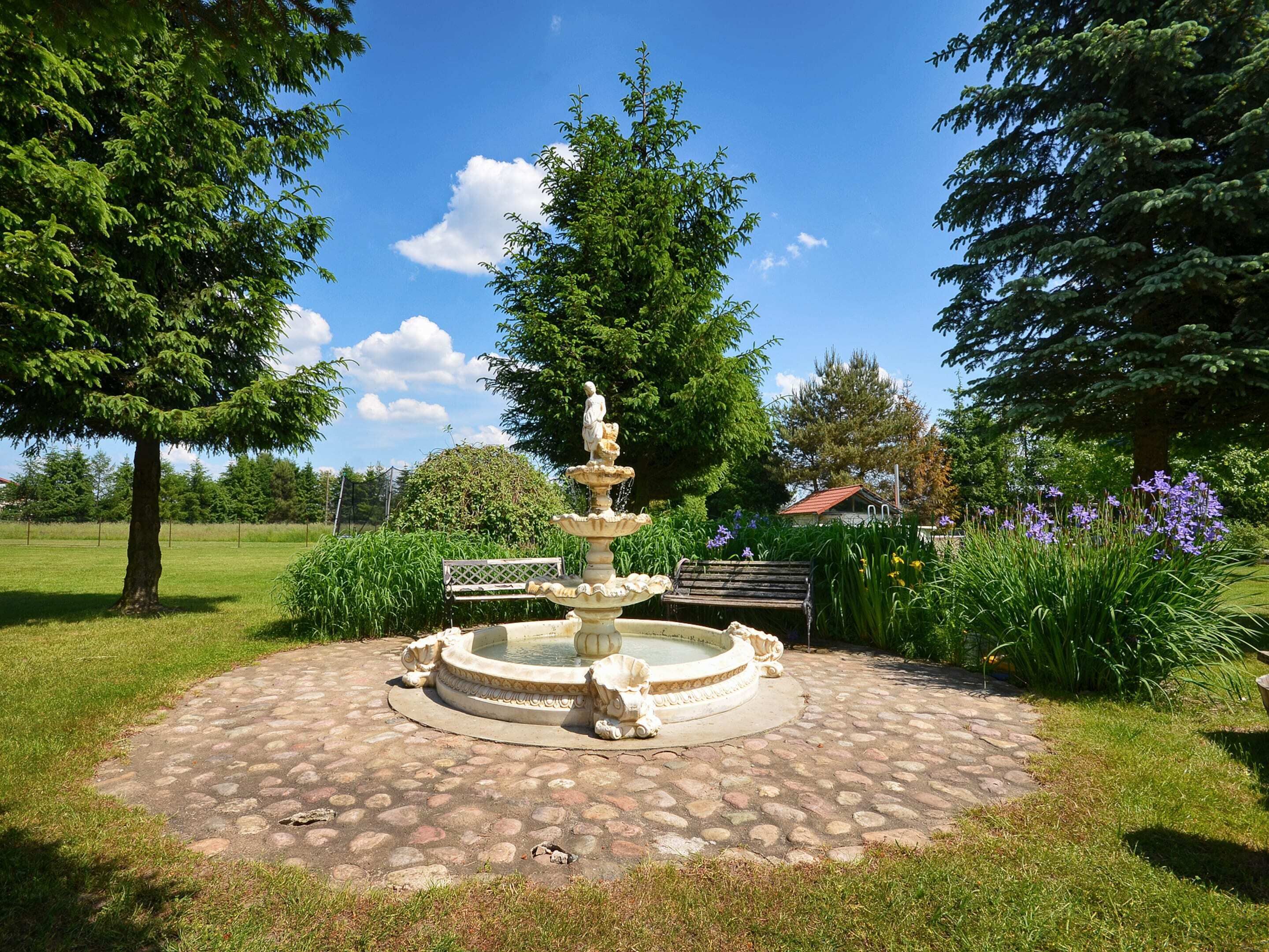 A circular stone fountain with a statue in the center, surrounded by a paved area with benches and lush greenery.