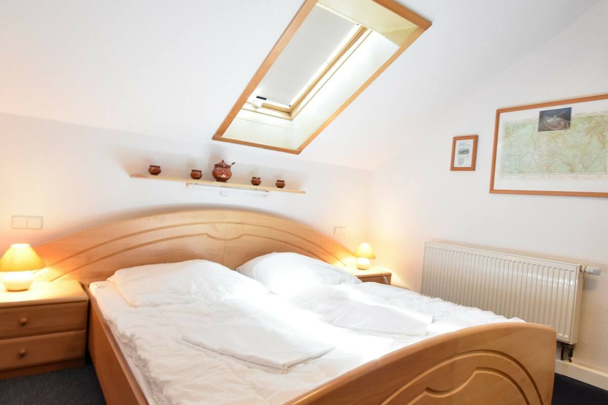 A neatly made bed in a hotel room with a wooden headboard, a skylight, bedside lamps, a radiator, and a shelf with decorative items.
