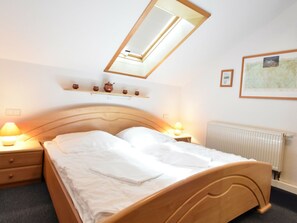 Een net opgemaakt bed in een hotelkamer, met een houten hoofdbord, een dakraam, bedlampen, een radiator en een plank met decoratieve objecten.