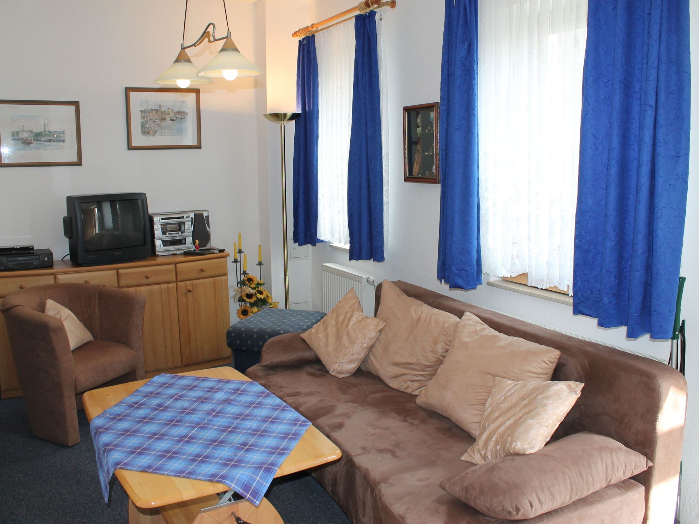 A living room with a brown sofa, a blue plaid blanket, a wooden coffee table, a television, and a small kitchenette.