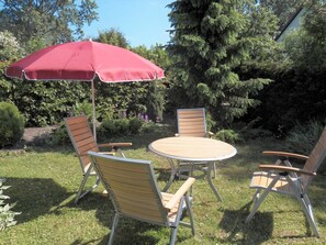 Een terras met een ronde tafel en vier stoelen, een rode parasol en een achtergrond van bomen en struiken.