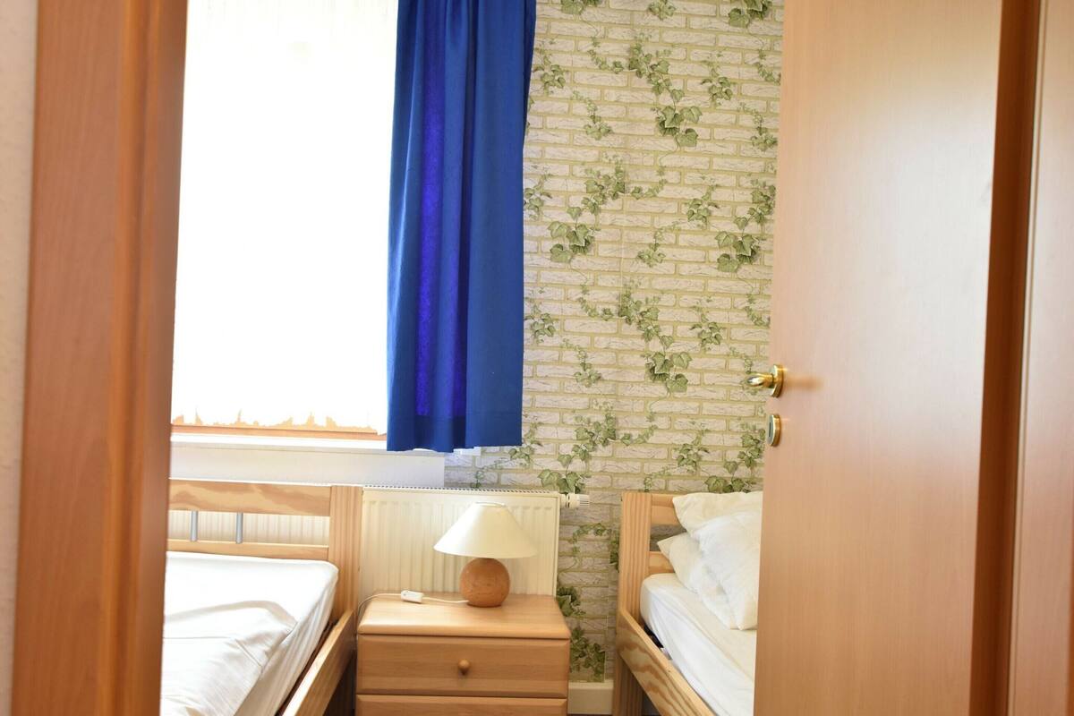 A small, well-lit room with two wooden beds, a nightstand with a lamp, and a brick-patterned wall.
