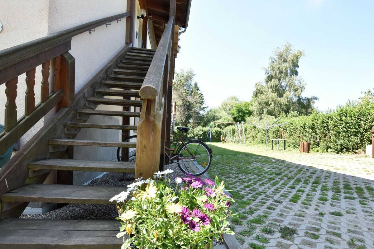 A wooden staircase leading to a building, a bicycle parked nearby, and a garden with flowers and a paved path.