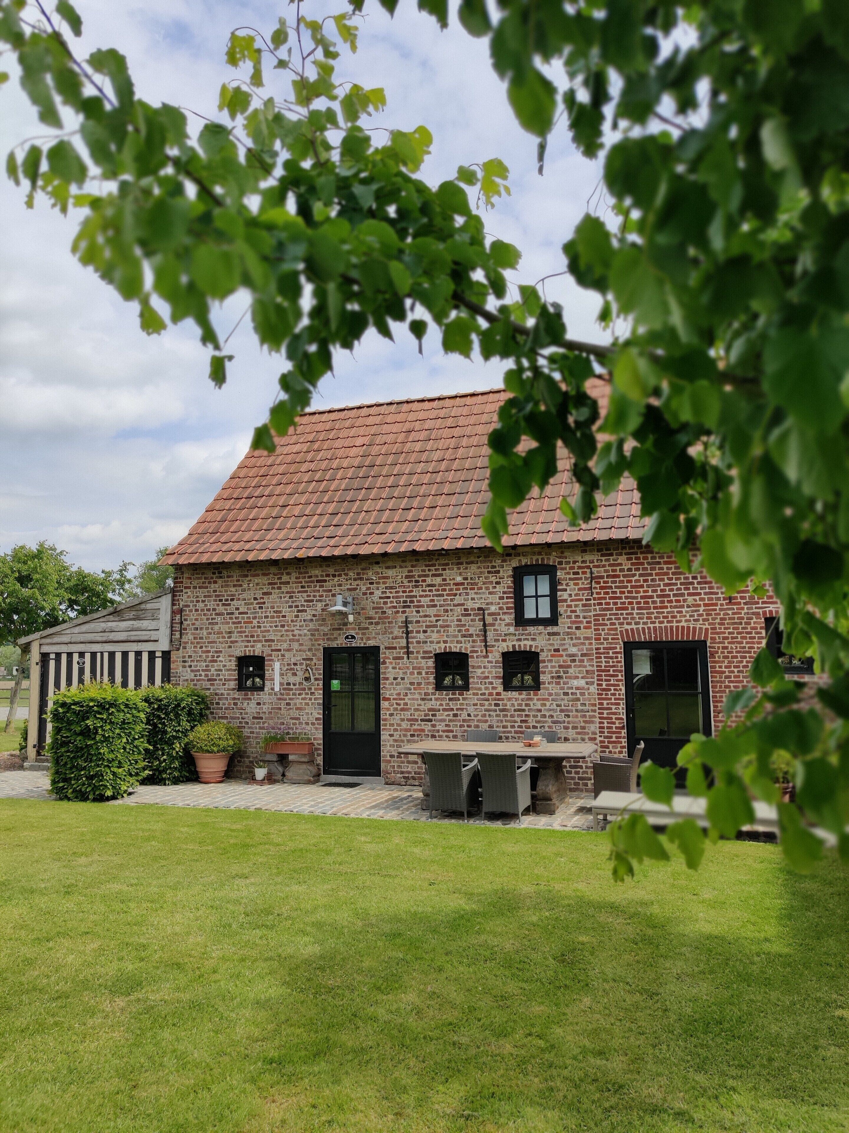 Een bakstenen gebouw met een pannendak, een zwarte deur en een kleine tuin met planten in potten.