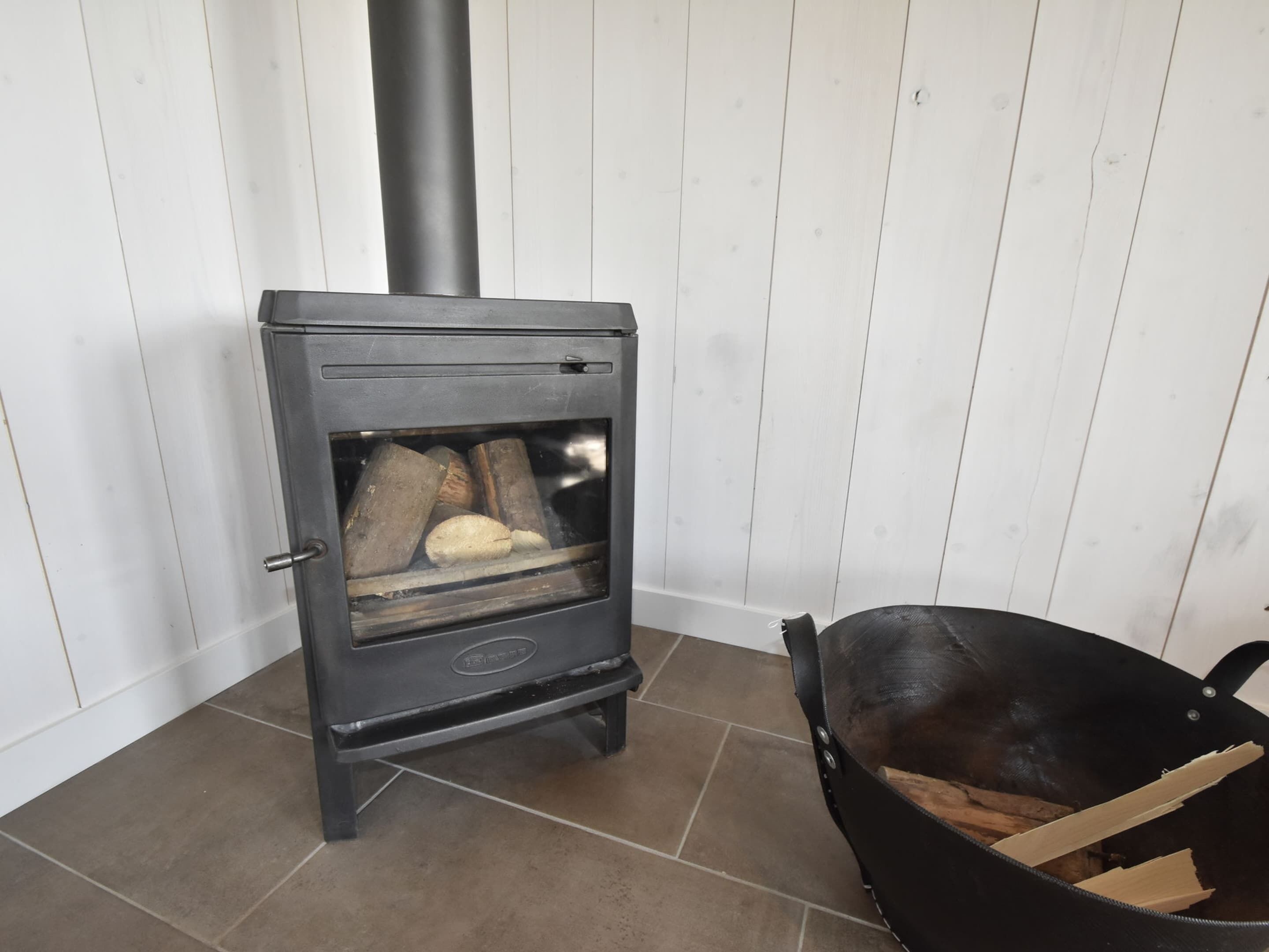 En vedeldad kamin med ved inuti, en svart metallkastrull på golvet och en vit trävägg.