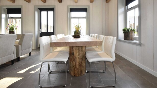 Un espace repas avec une table en bois entourée de chaises blanches, un plafond en bois et de grandes fenêtres.