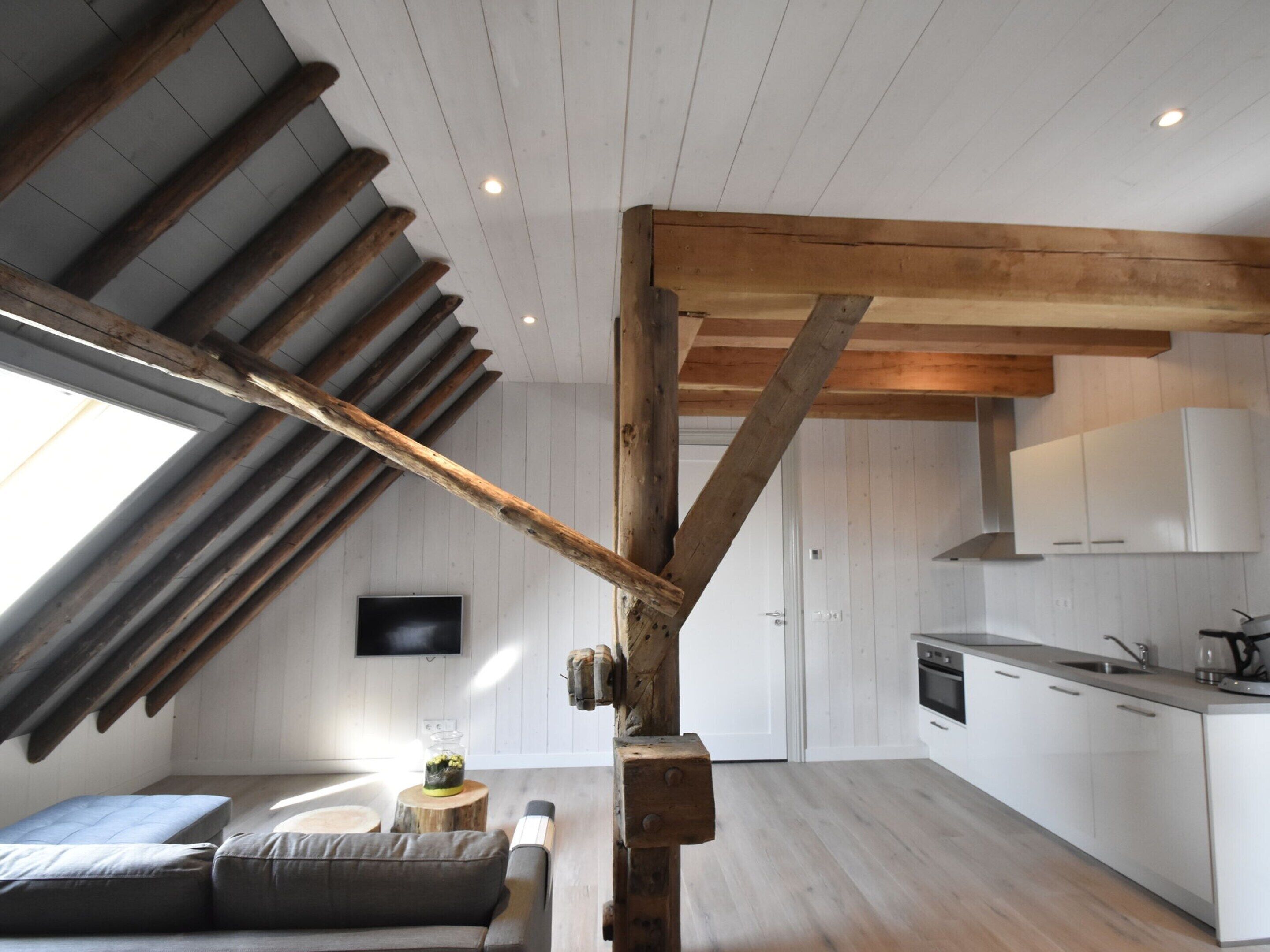 Ein Loft-Zimmer mit freiliegenden Holzbalken, einer integrierten Küche, einer Couch und einem großen Fenster.