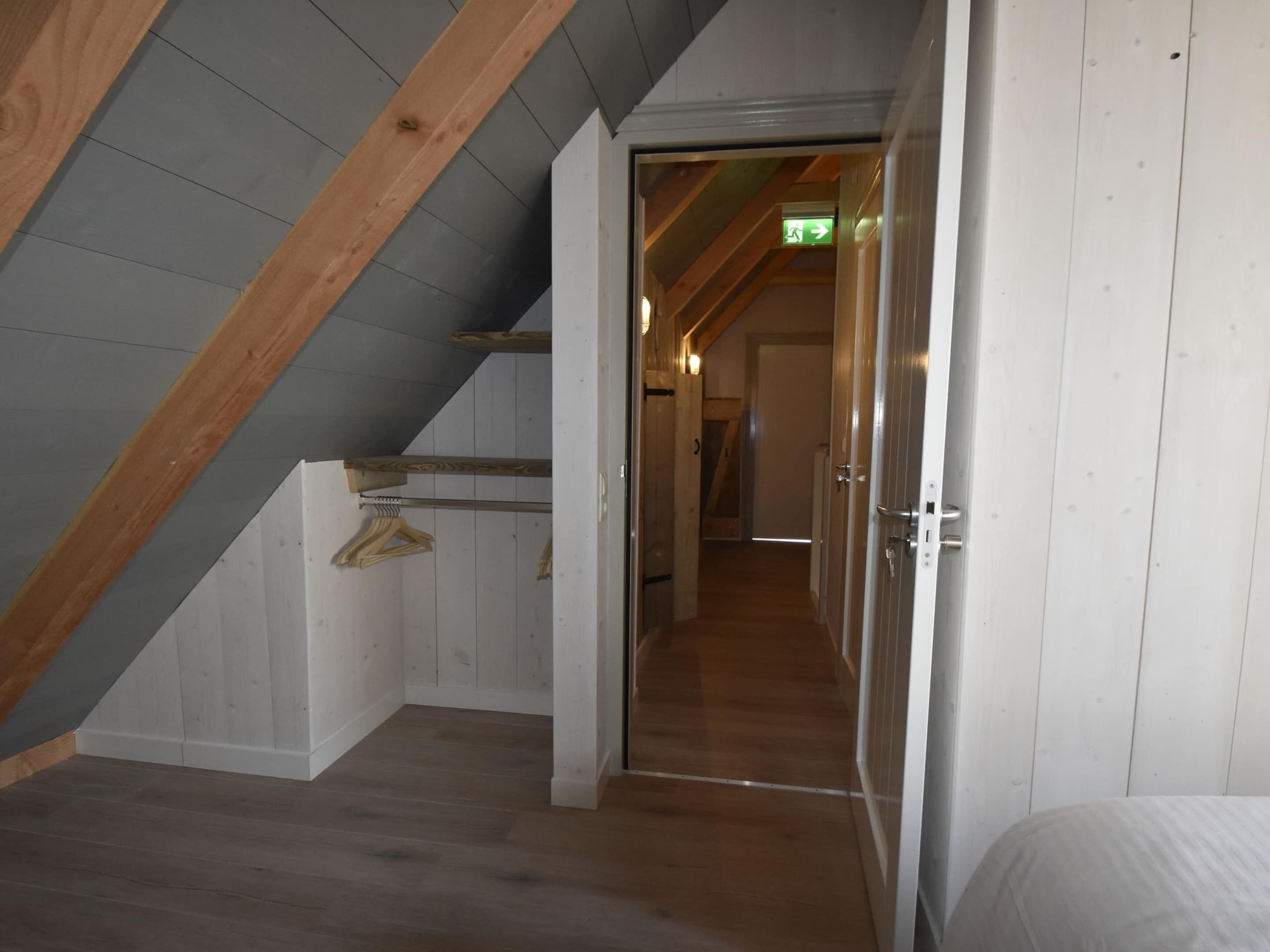 Un couloir étroit aux poutres apparentes et au plafond en pente, qui mène à une pièce contenant un lit et une porte.