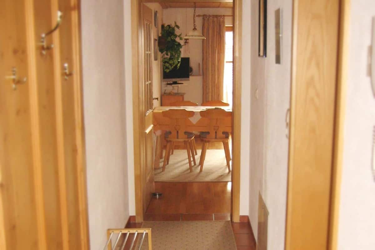 Een smalle gang die uitkomt in een eetruimte met een houten tafel, stoelen, een televisie en een plant.