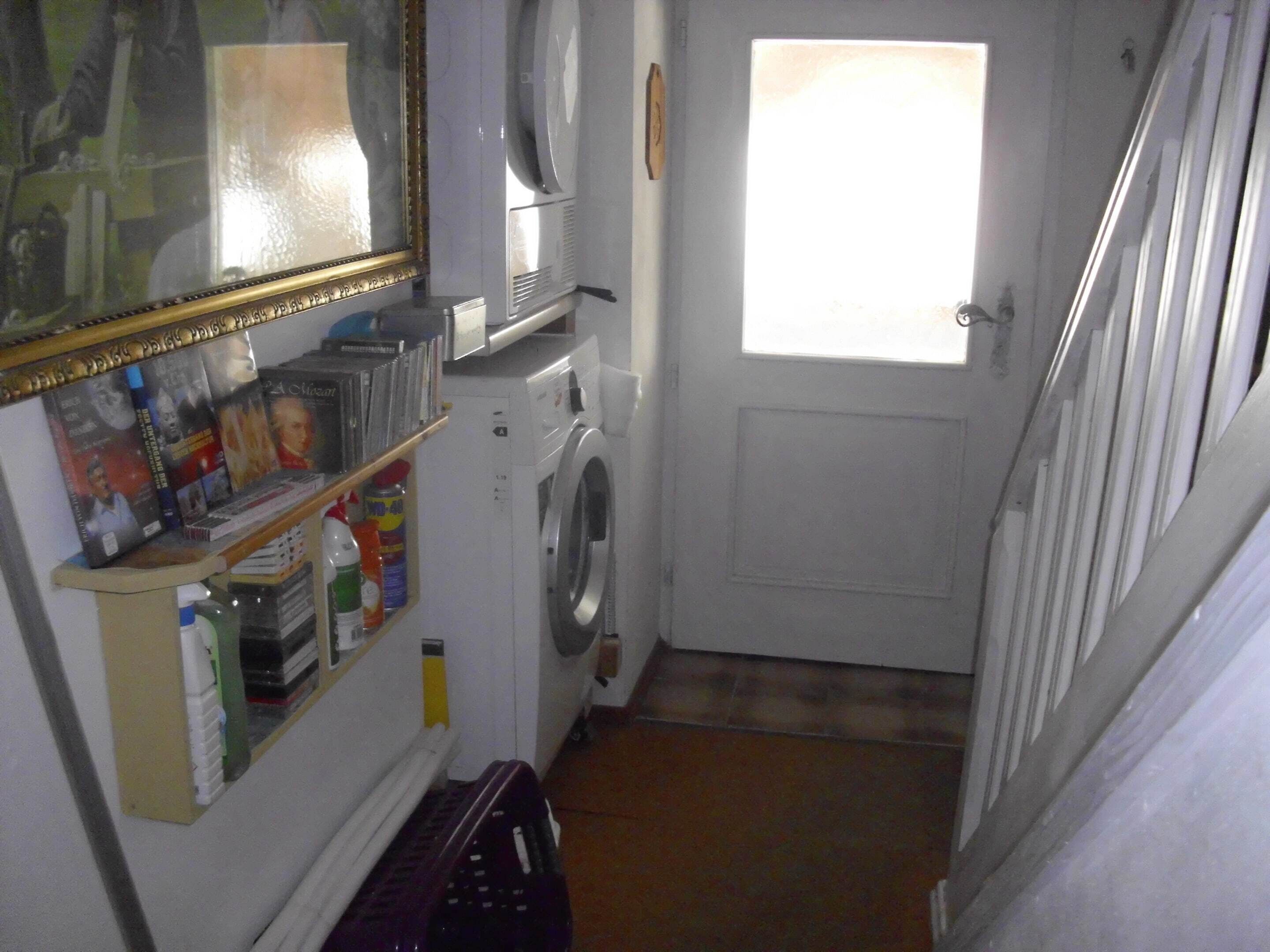 A laundry room with a washing machine, a shelf with books and cleaning supplies, and a door leading to another room.