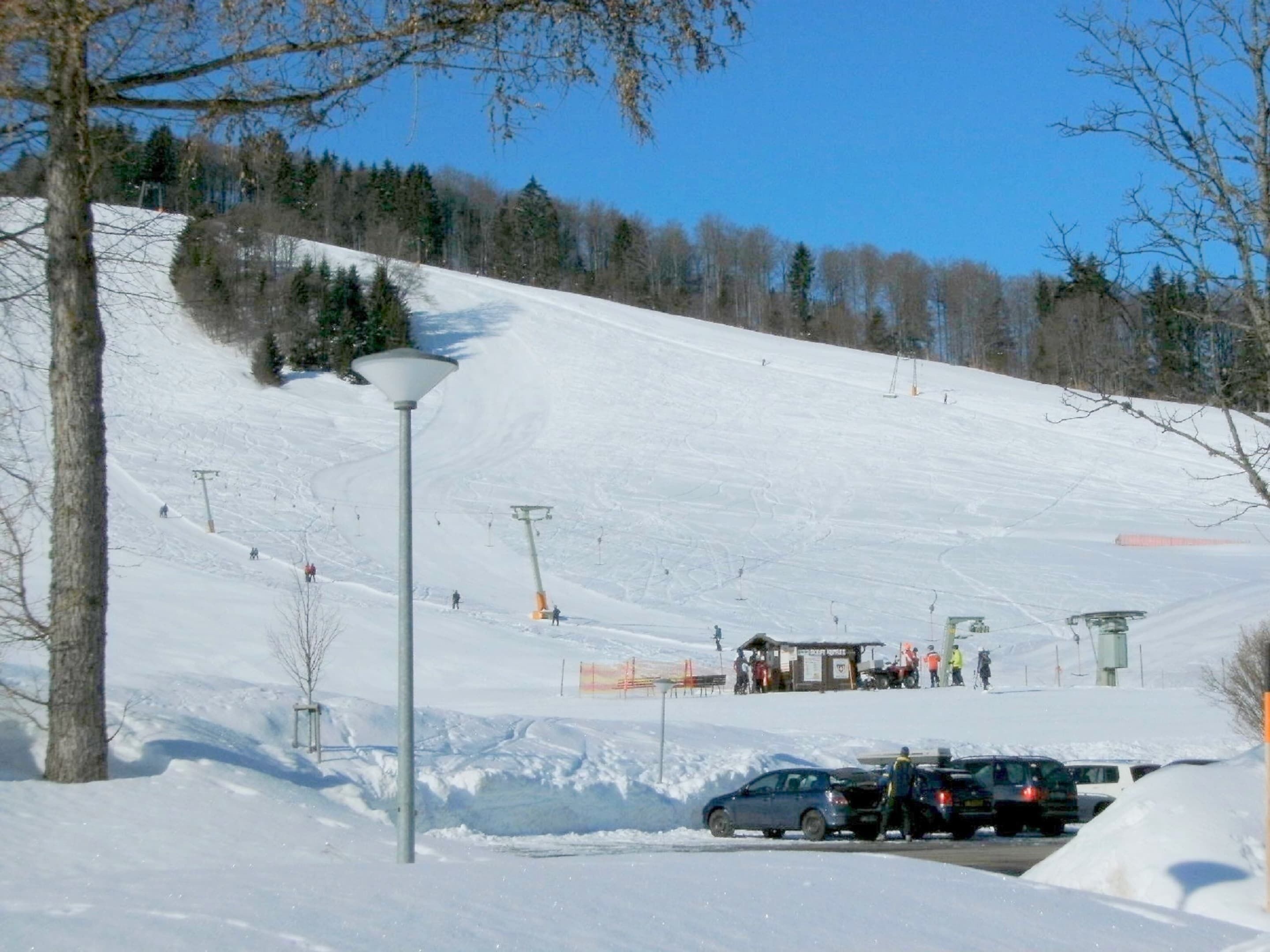 Ein verschneiter Skipisten mit Sessellift, Parkplatz und einem kleinen Gebäude.