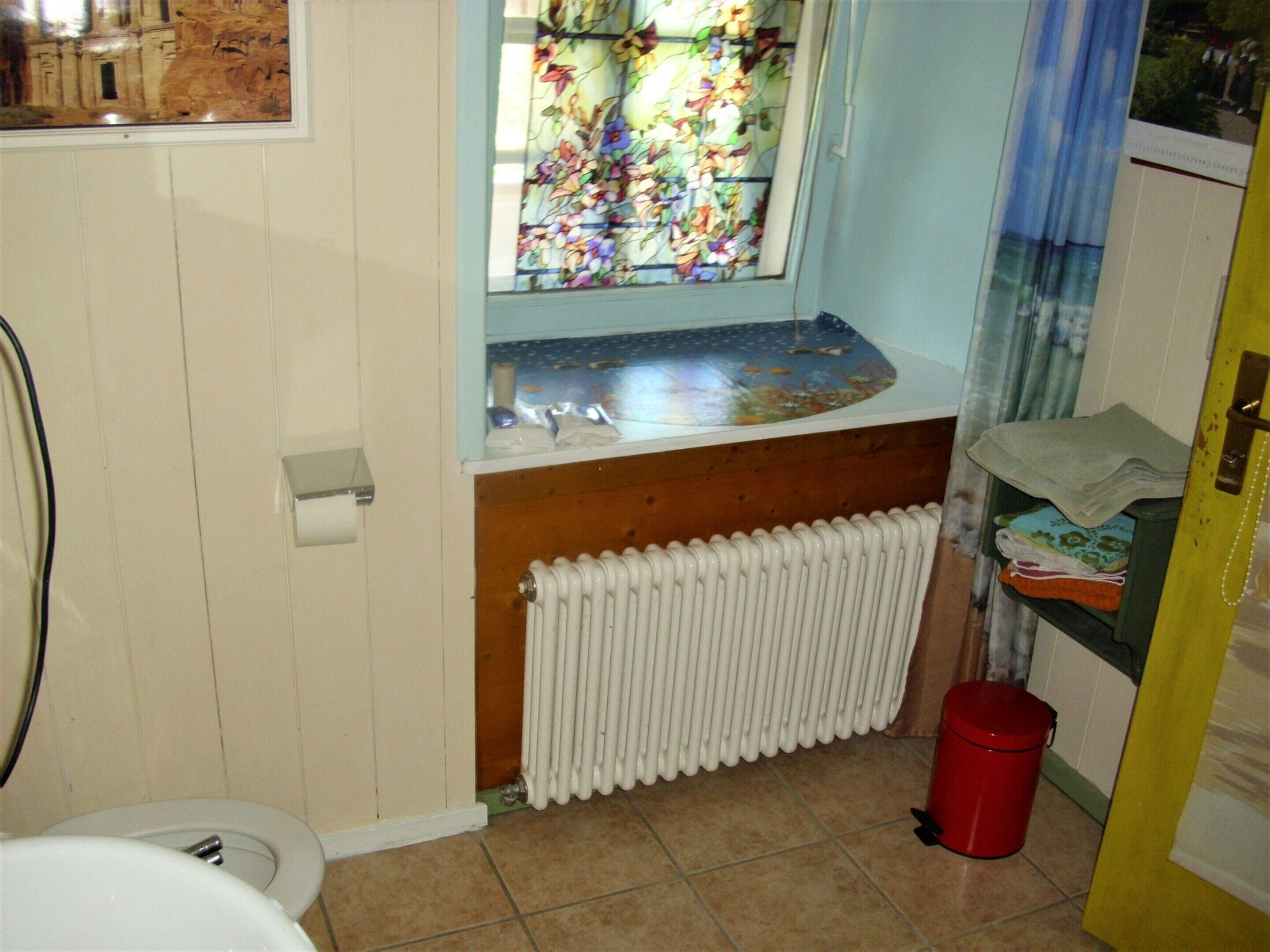 A bathroom with a toilet, a window with a floral stained glass design, a radiator, and a red trash bin.