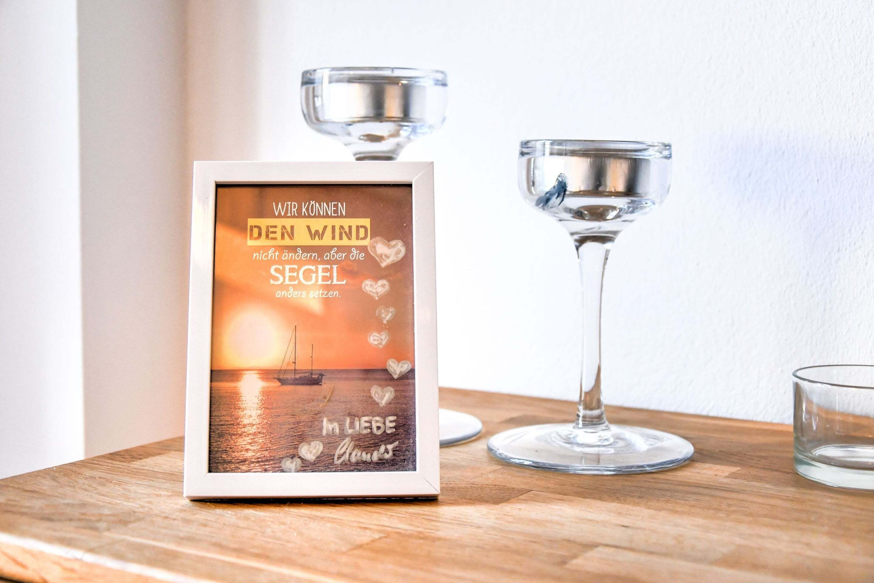 Une citation encadrée avec une vue sur un coucher de soleil, deux verres en verre transparent et une surface en bois.