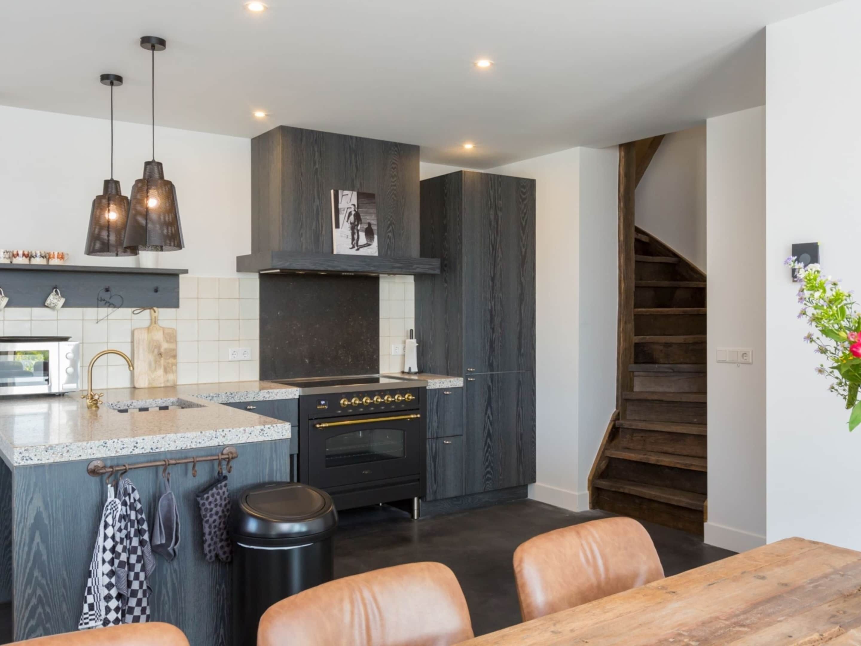 A modern kitchen with a central island, dark wood cabinetry, and a built-in oven. There is a dining area with leather chairs and a wooden table.