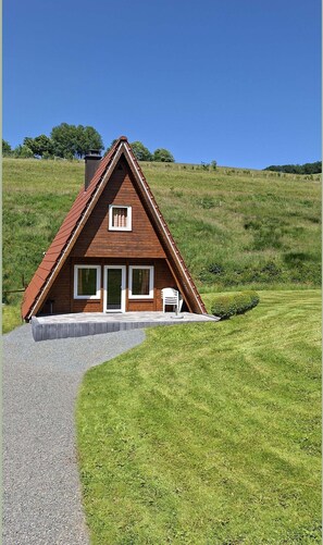 A wooden house with a sloped roof, a paved entrance, and a small patio with a chair. - Cosy Holiday Home in the Hochsauerland With Terrace at the Edge of the Forest (Bestwig)