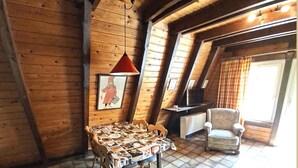 Une pièce mansardée chaleureuse avec un plafond en bois, une table à manger recouverte d’une nappe à motifs, une seule chaise, un téléviseur et une fenêtre avec des rideaux.
