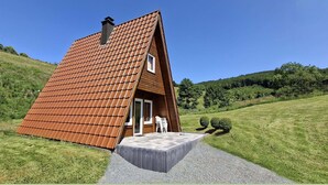 Une petite cabane en bois, dotée d’un toit en pente, d’un porche avec un banc et offrant une vue sur une colline verdoyante.