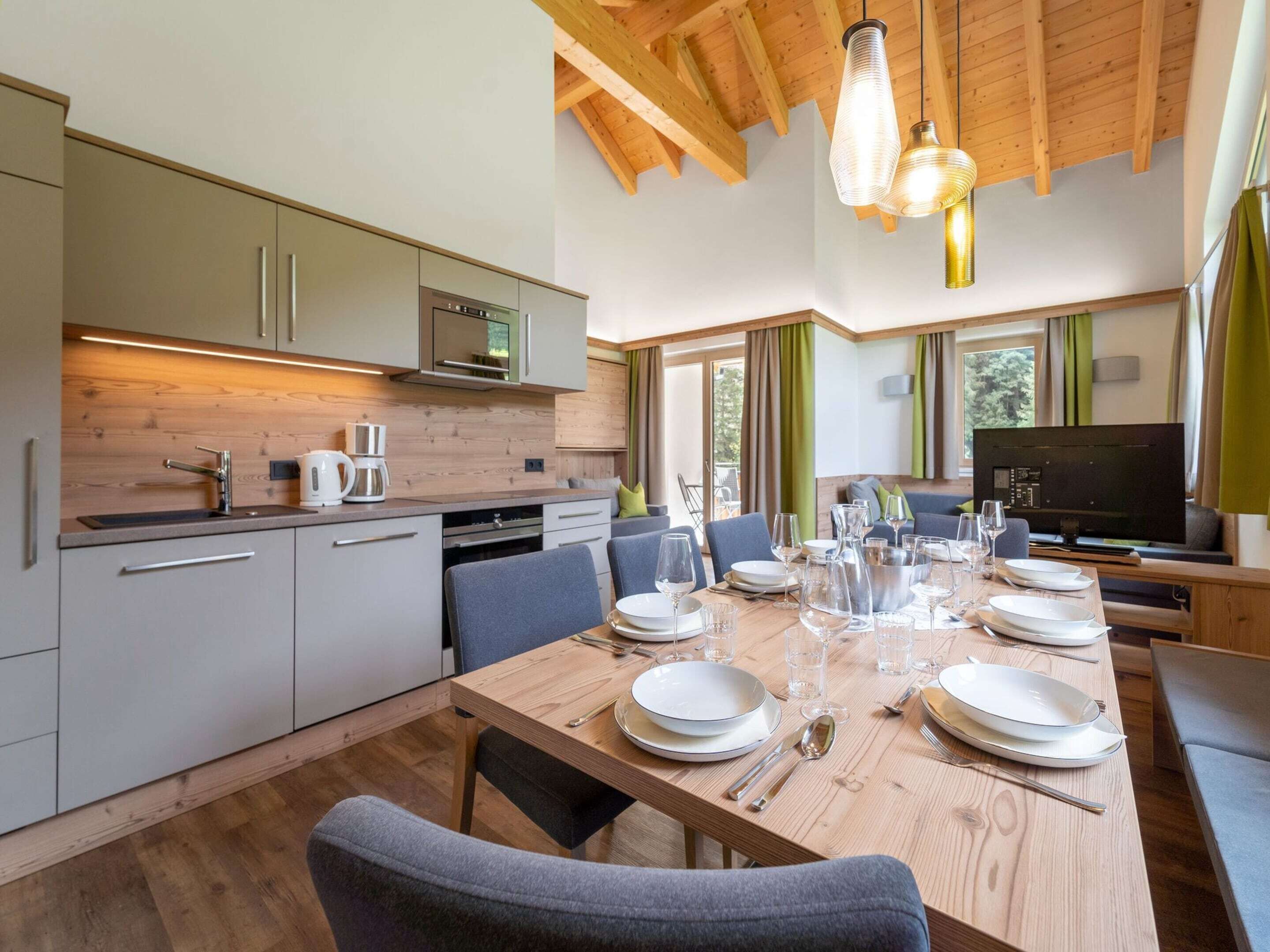 A modern kitchen with wooden beams, a dining area with a long table set for six, and a TV in the background.