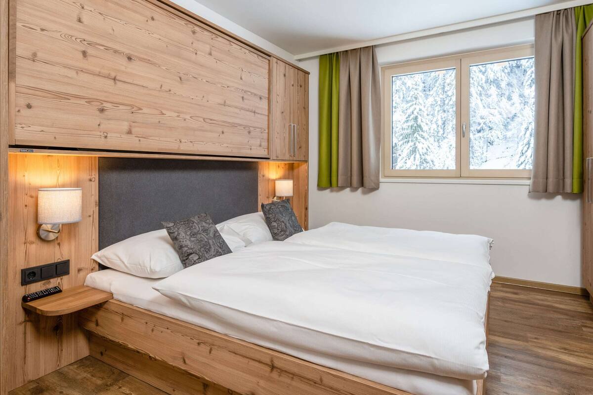 A bedroom with a wooden headboard, a bed with white bedding, a nightstand with a lamp, and a window with curtains.