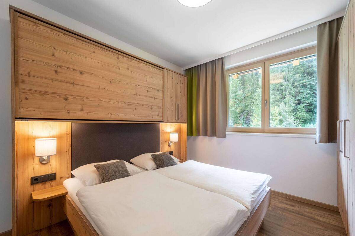 A double bed with a wooden headboard and nightstands, a window with curtains, and a view of trees.