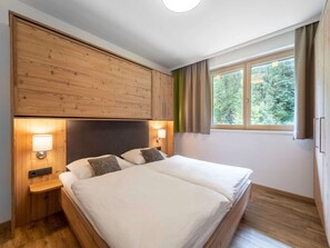 Ein Doppelbett mit Holzüberbau und Nachttischen, ein Fenster mit Vorhängen und Blick auf Bäume.