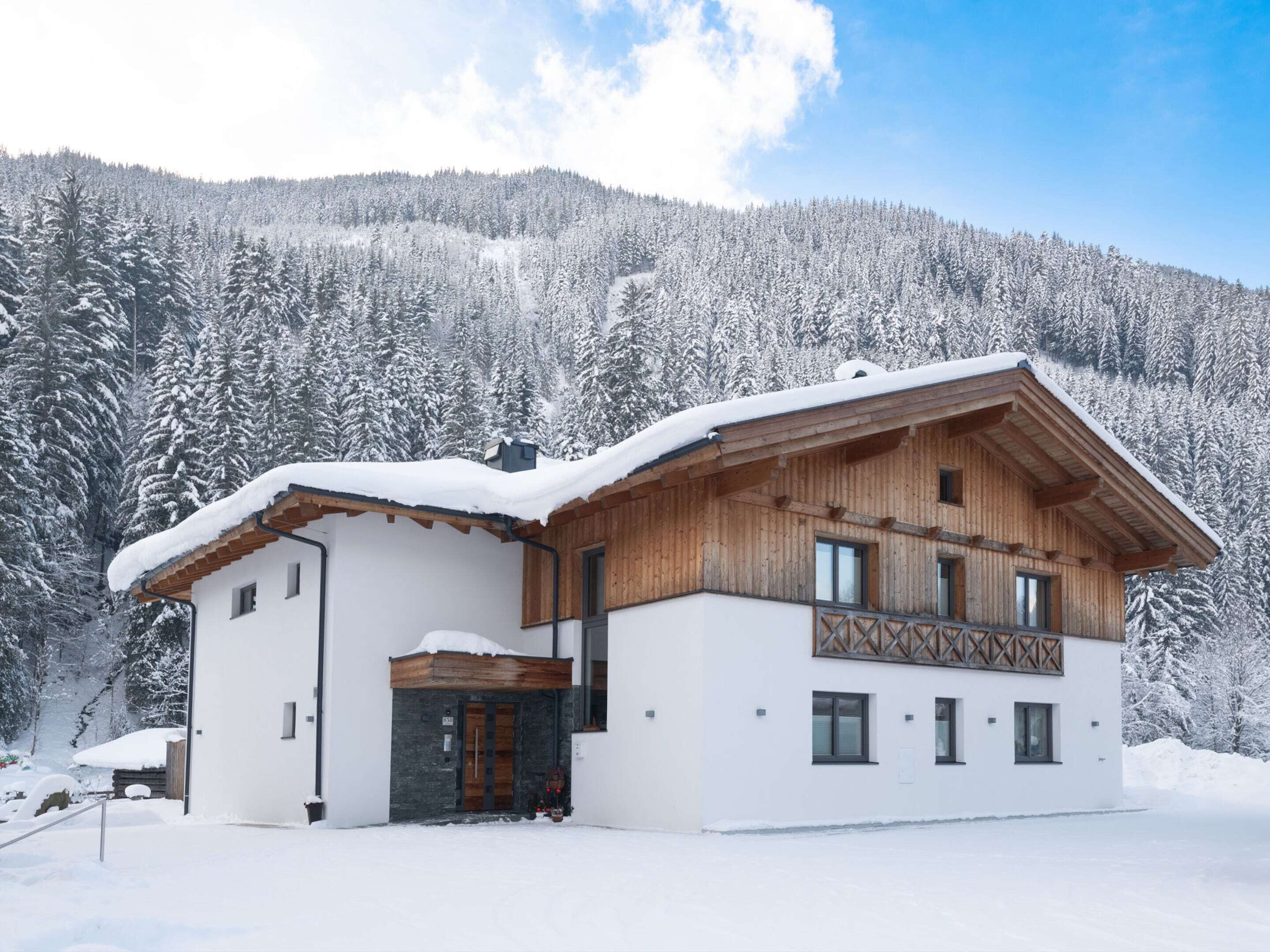 A modern chalet with a wooden facade, white exterior walls, and a snow-covered roof, set against a snowy forest backdrop.