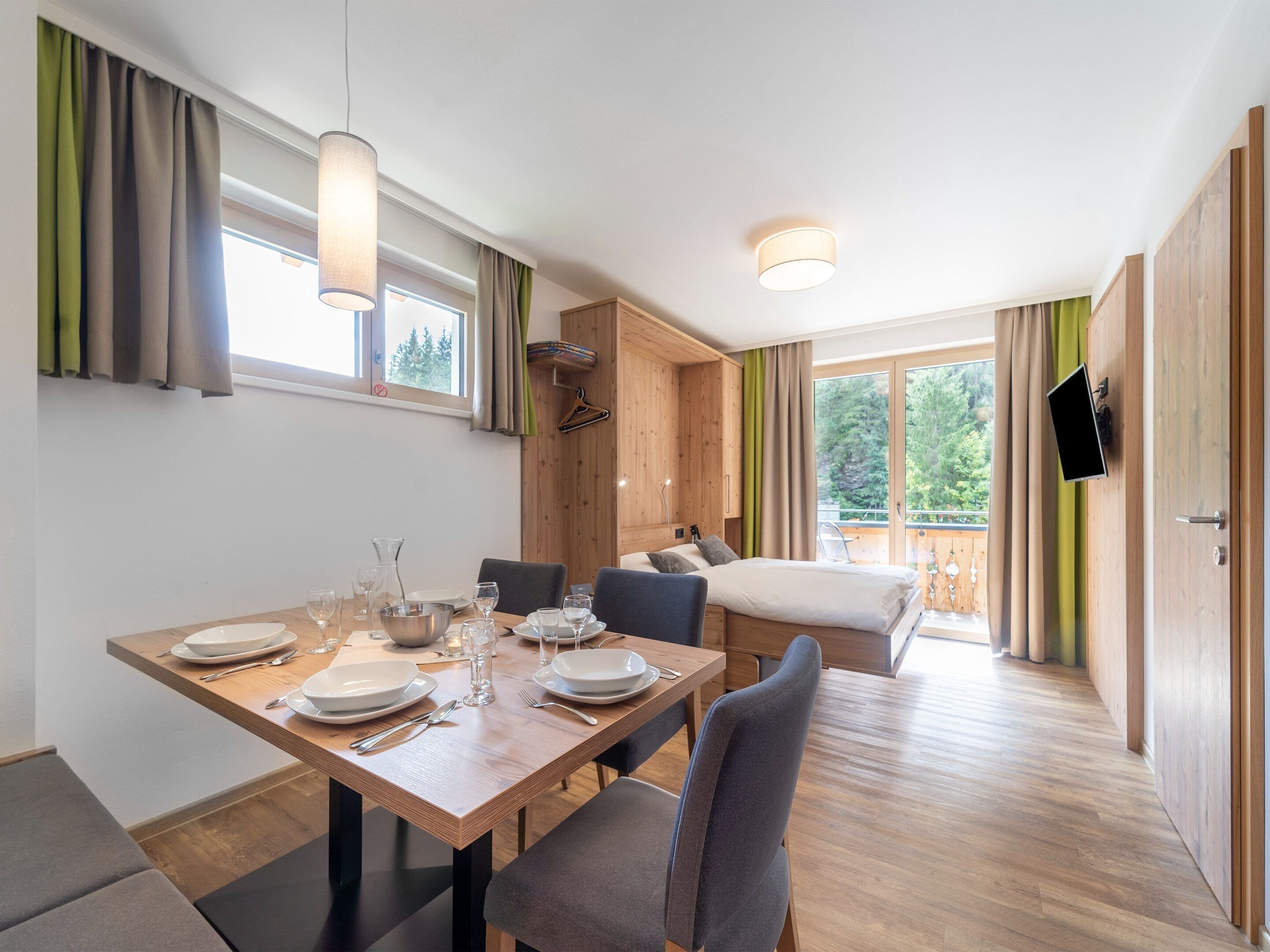Une chambre moderne avec une table à manger en bois, préparée pour quatre personnes, un lit avec vue et un téléviseur fixé au mur.