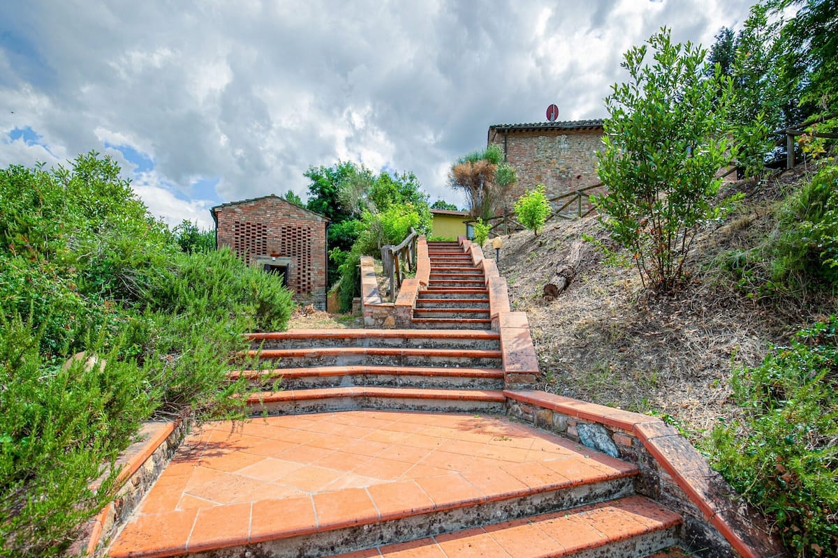 A set of terracotta-tiled stairs leading up a hill, surrounded by greenery and a rustic building in the background.