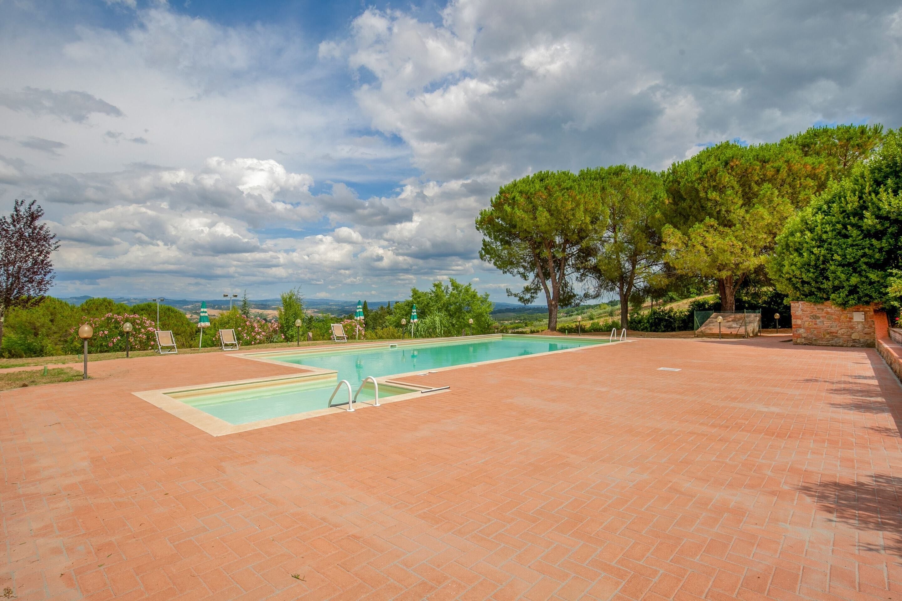 A rectangular swimming pool with a tiled deck, surrounded by trees and a stone wall.