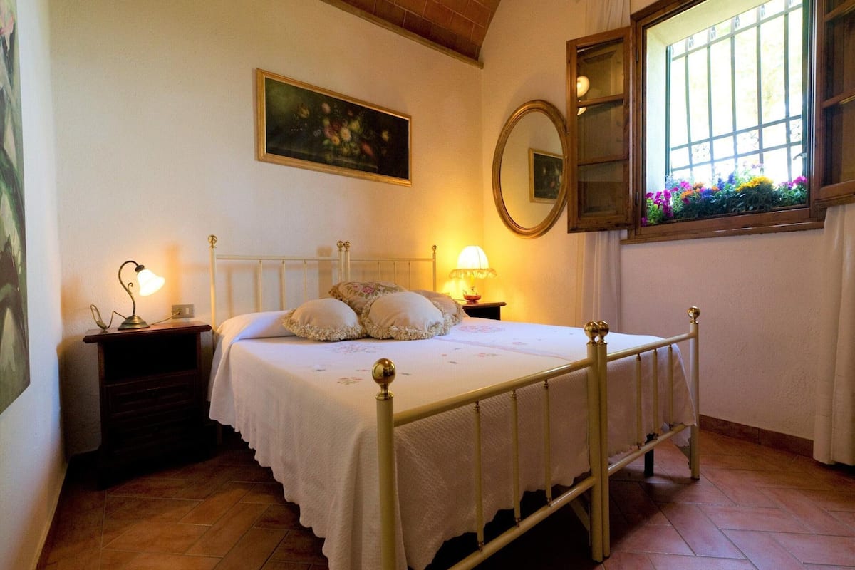 A bedroom with a bed, nightstands, a mirror, a window with flowers, and a painting on the wall.