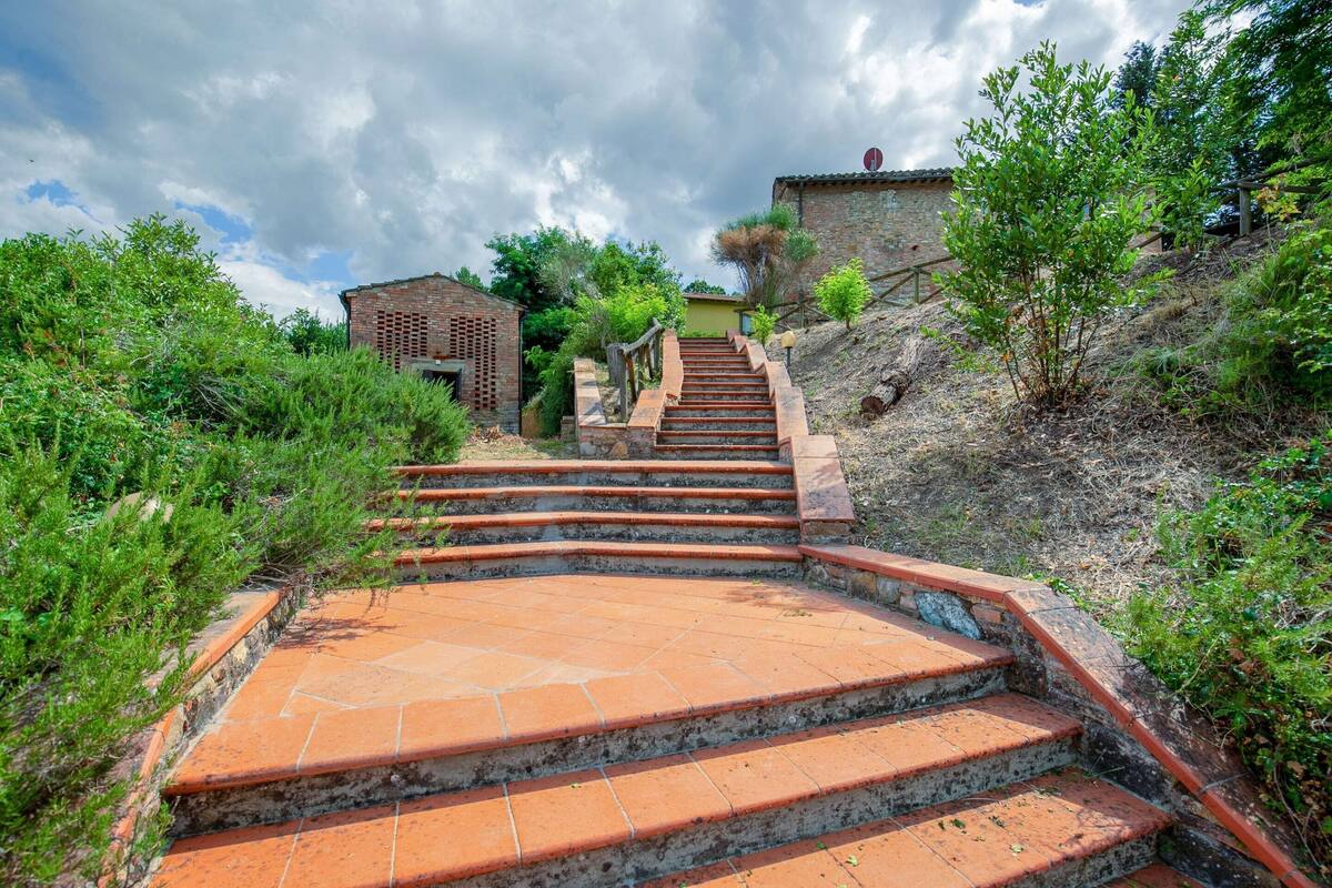 A set of terracotta brick stairs leading up a hill, surrounded by greenery and a rustic building in the background.